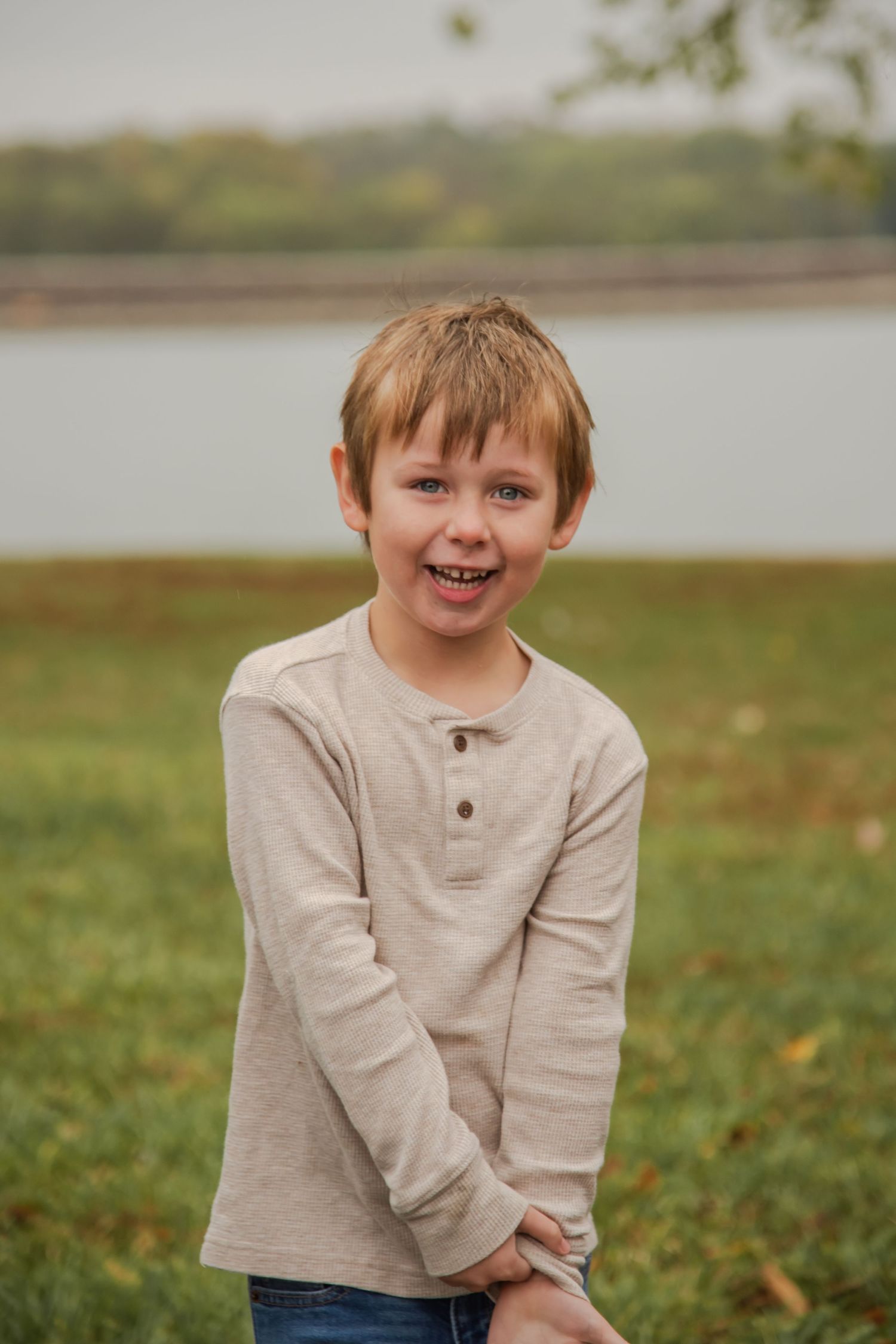 A child in a beige henley shirt smiles playfully while standing in a grassy field on an autumn day.