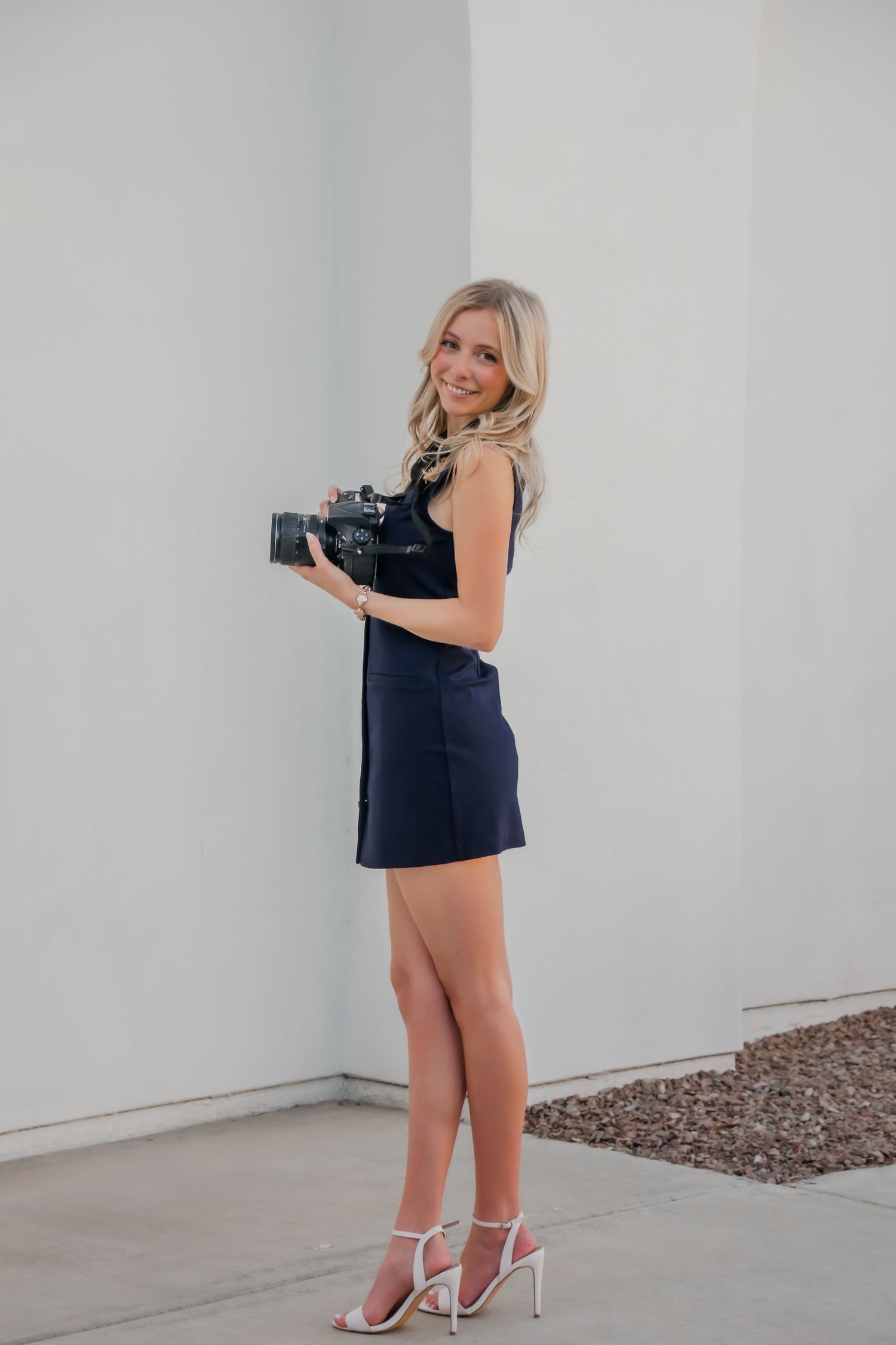 Blonde woman in navy mini dress and white heels holding a camera, posing against a light gray wall outdoors.