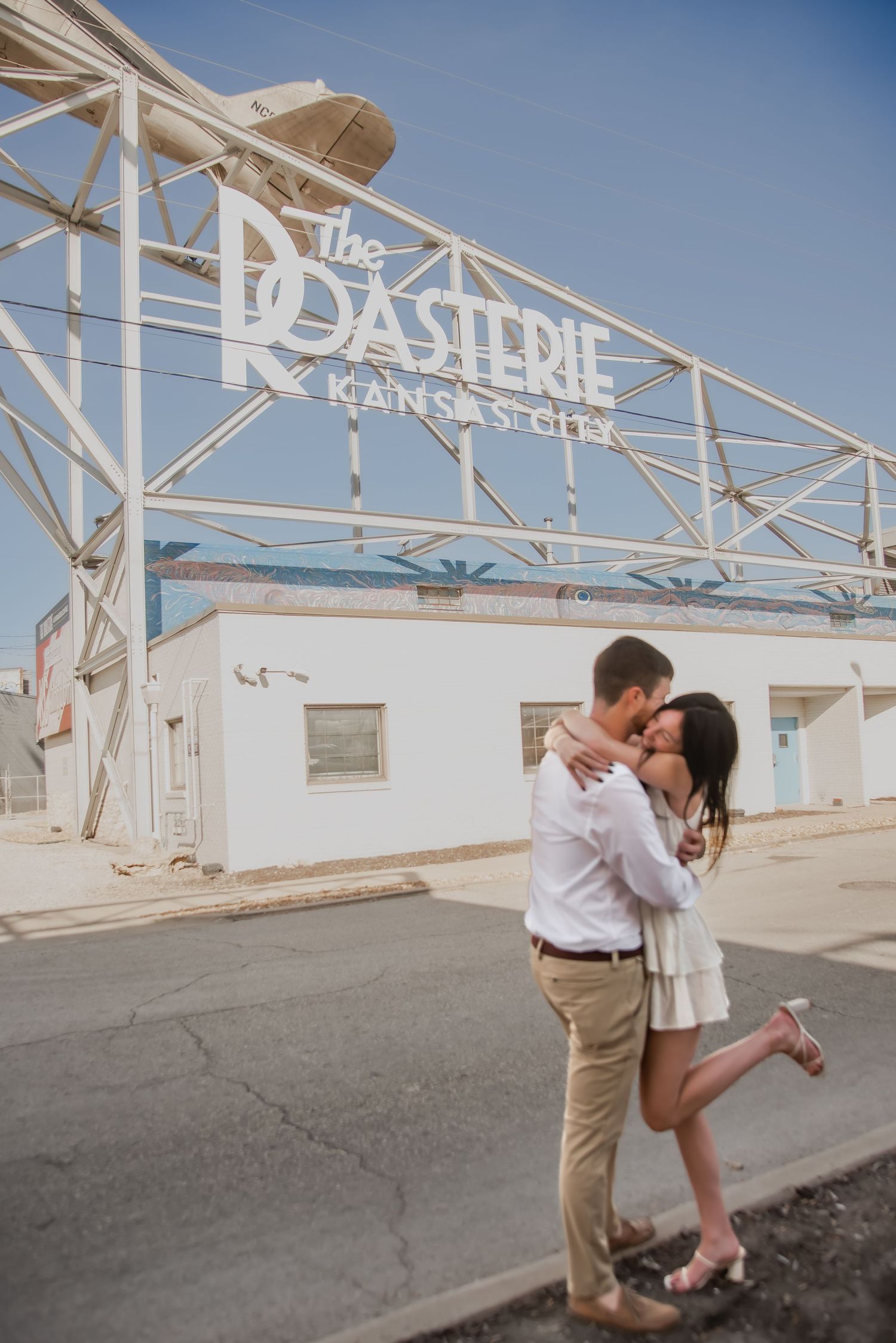 A couple shares a romantic moment in front of a white industrial building with architectural framework details.