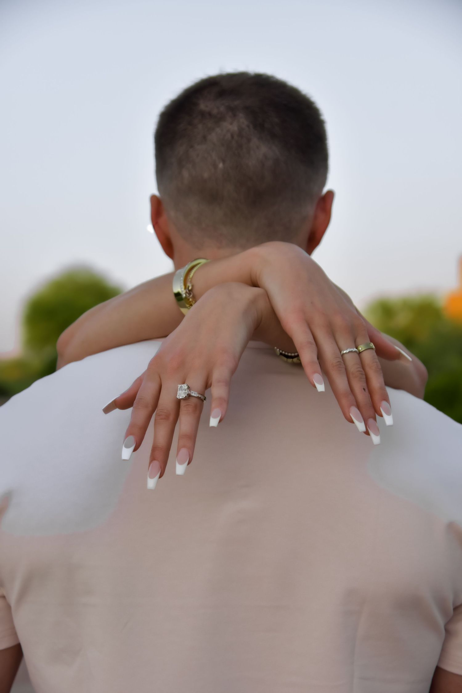 Close-up shot of manicured hands with engagement ring embracing shoulders in romantic outdoor setting.