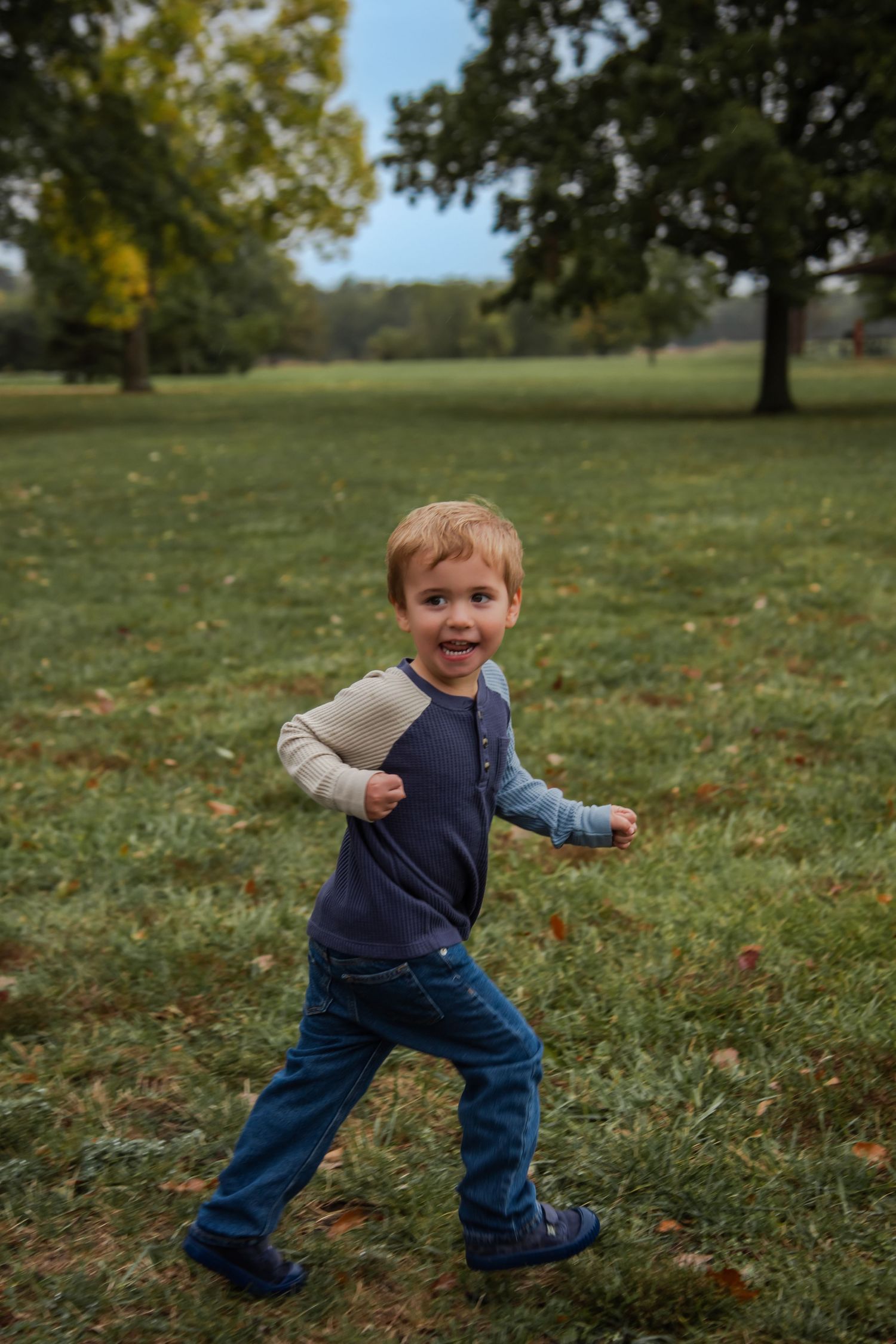 A small child in blue jeans and a navy shirt runs playfully through a grassy park with autumn leaves scattered about.