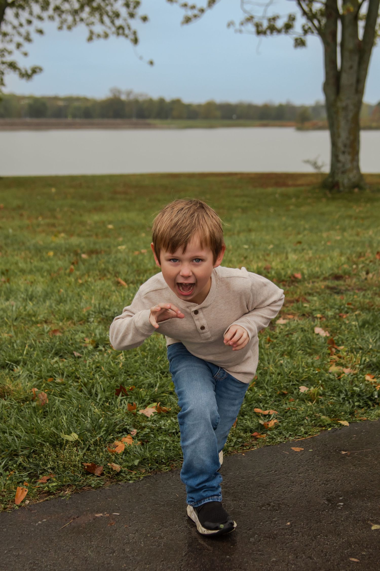 A child in a beige sweater and jeans runs playfully through fallen autumn leaves on a grassy field.