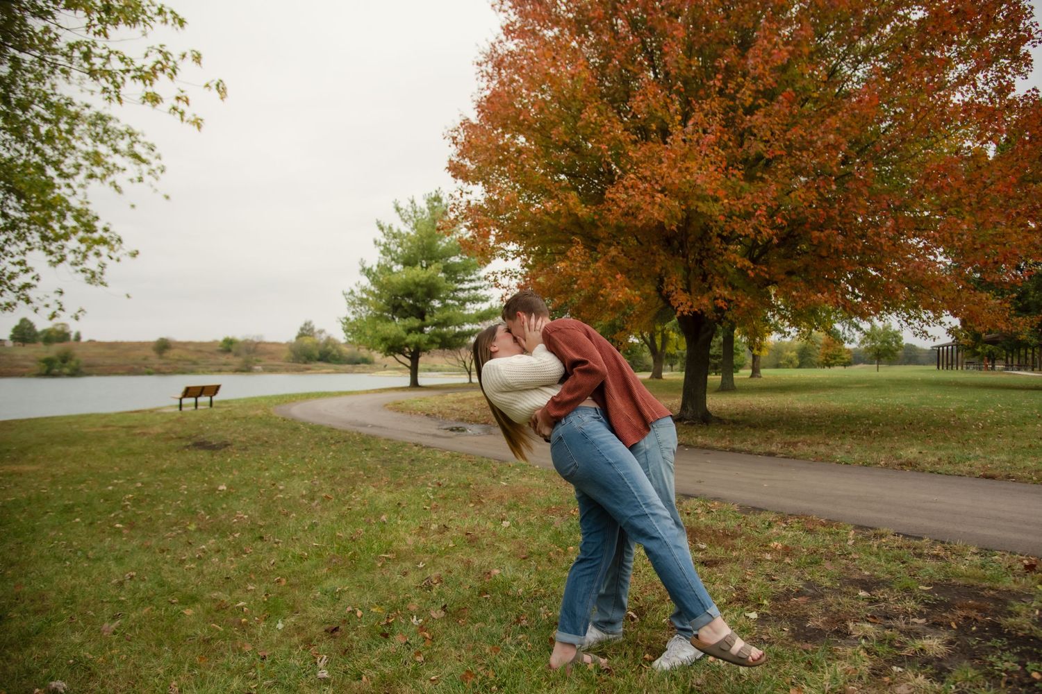 A couple embraces on an autumn day near a lake with fall foliage and a dog in the background.
