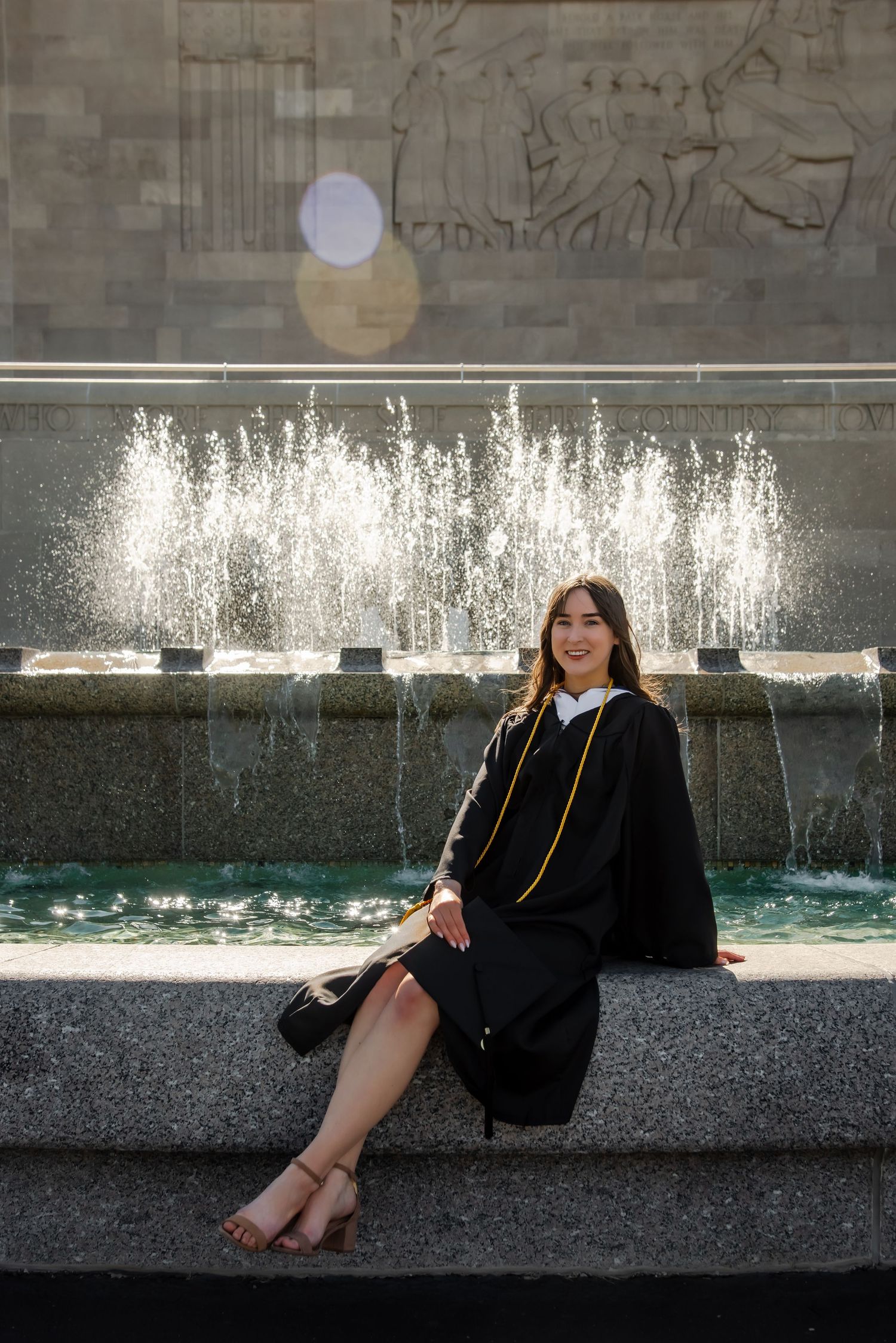 Graduate posing in front of a cascading water fountain wearing a black graduation gown.