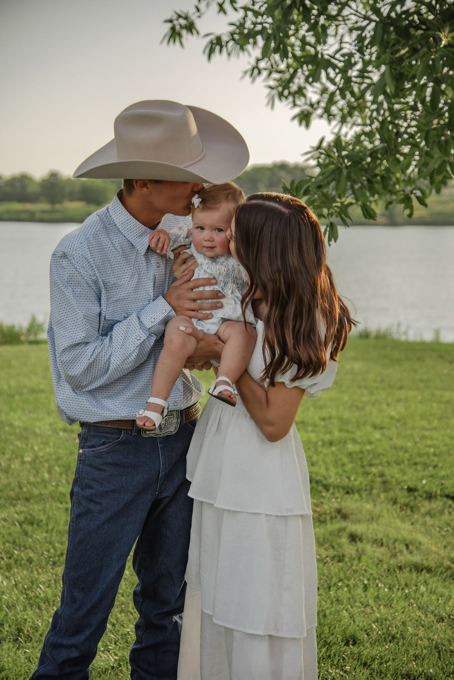 A tender family moment by a lake with a western-style cowboy hat and flowing white dress at sunset.