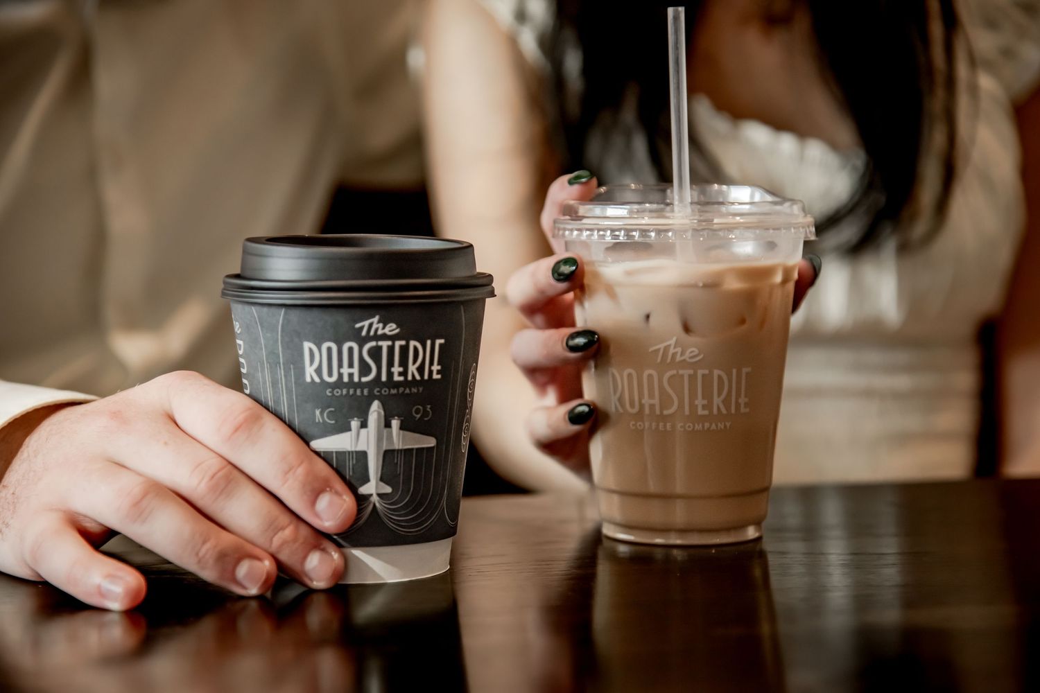 Multiple hands holding coffee cups and beverages from Roast & Serve cafe in a warm, cozy setting.