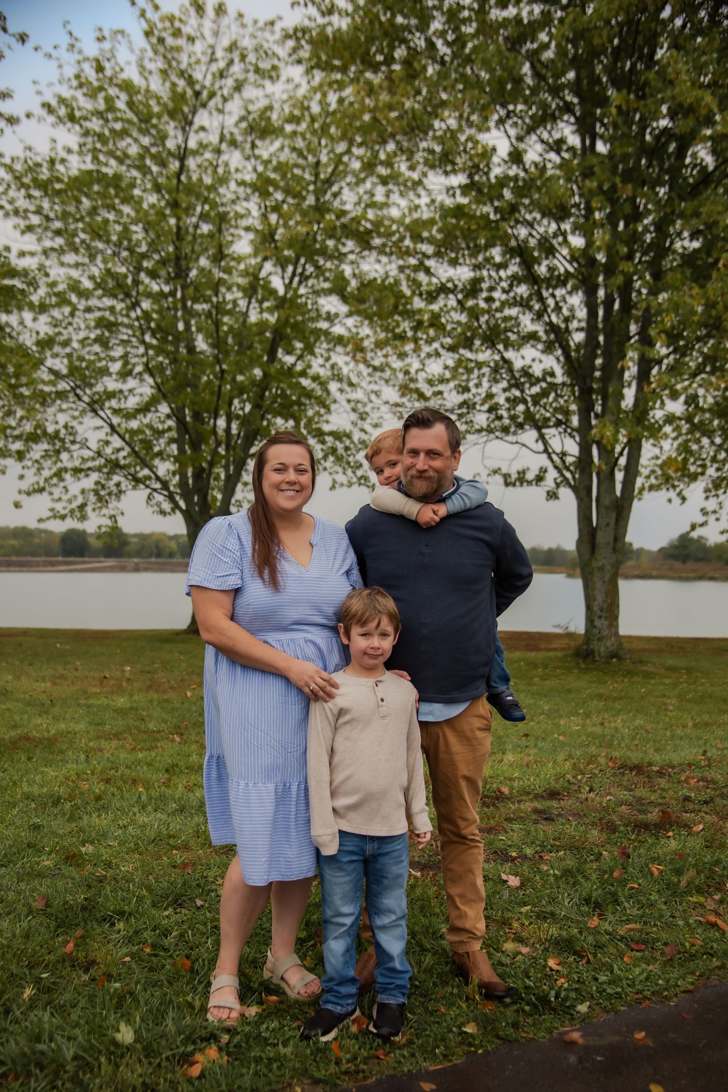 A family stands together on grassy terrain near trees and a lake on an autumn day.