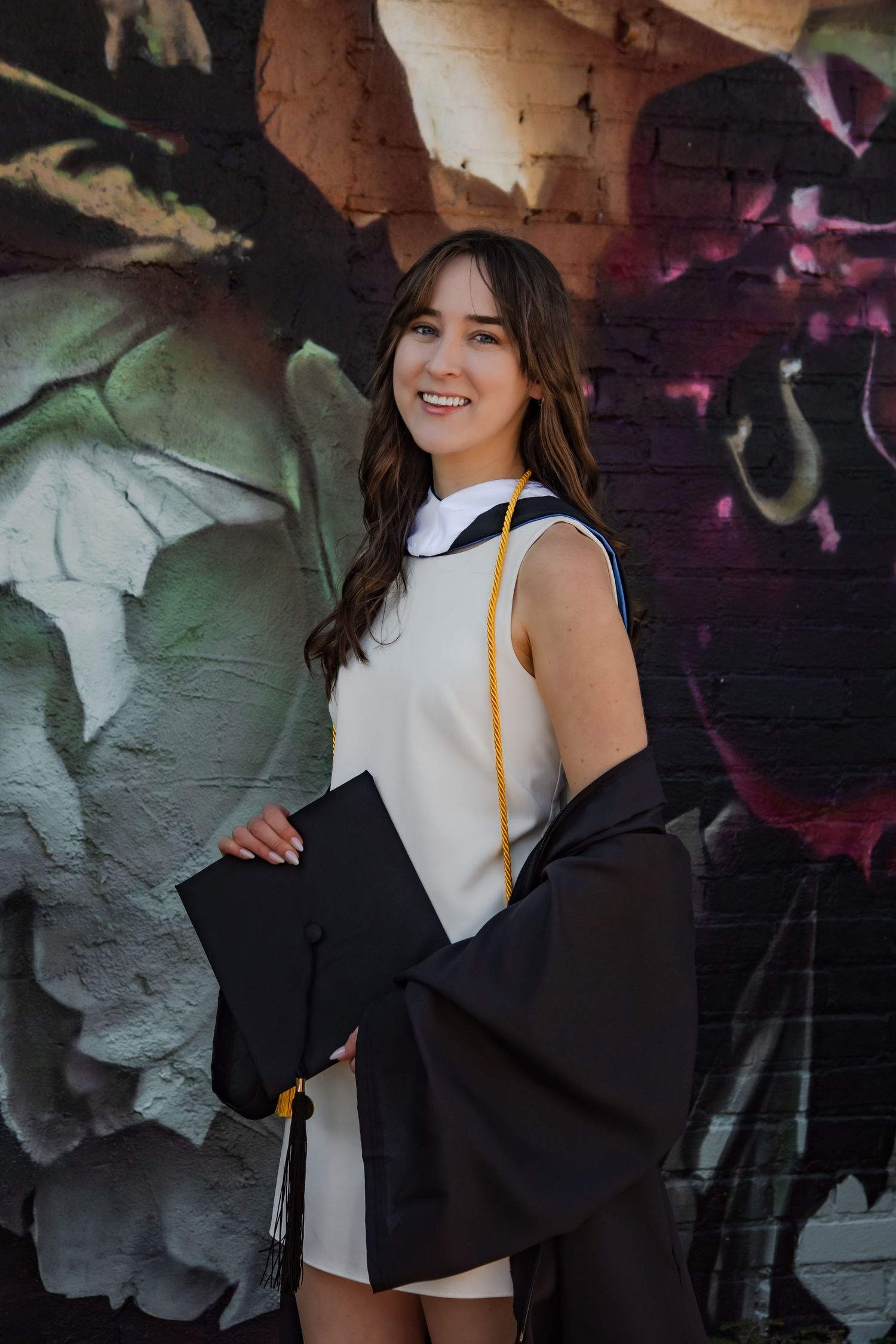 Graduate posing in white dress and black gown against colorful graffiti wall.