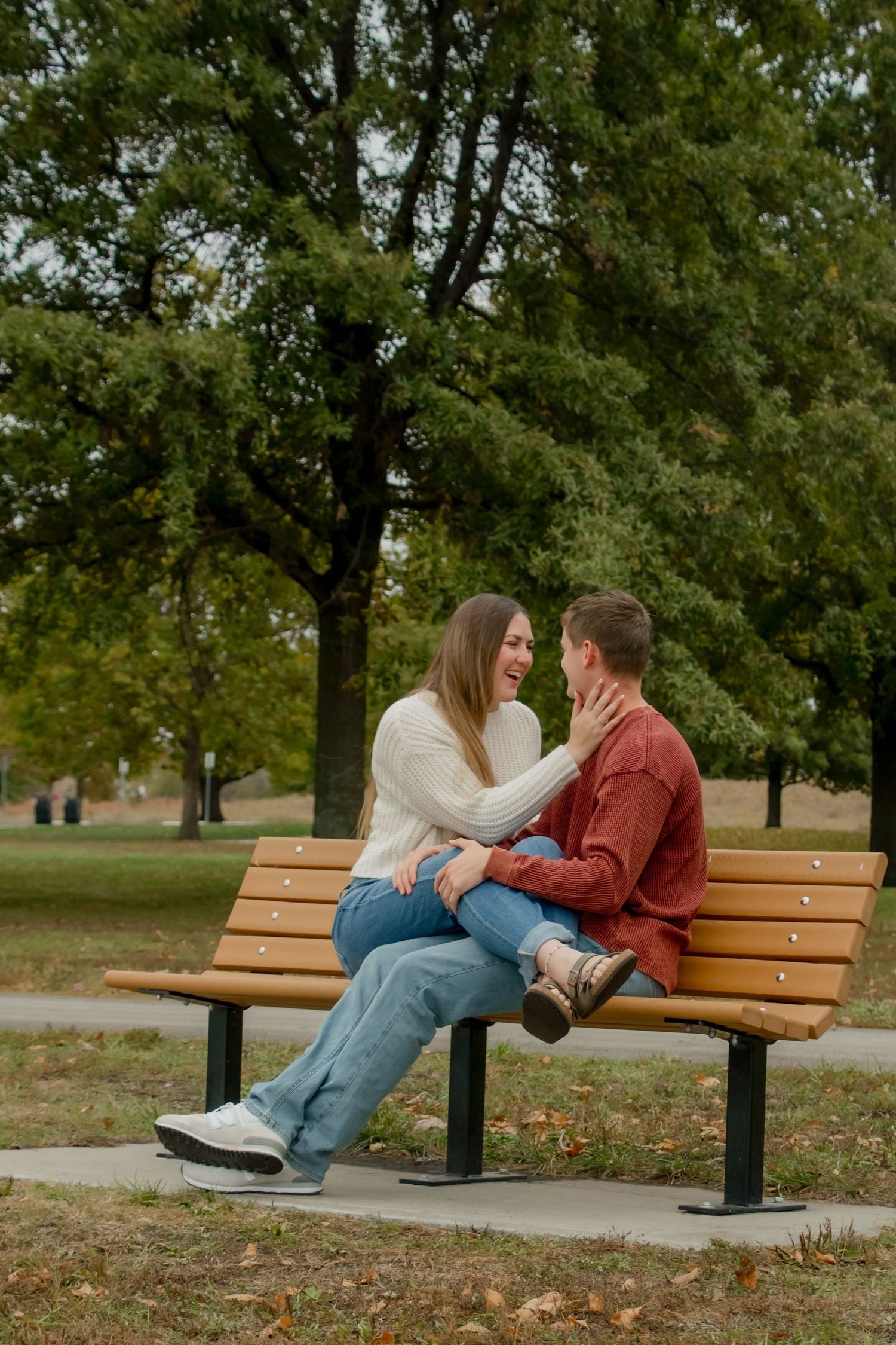 A couple sits close together on a wooden park bench surrounded by autumn trees and fallen leaves.