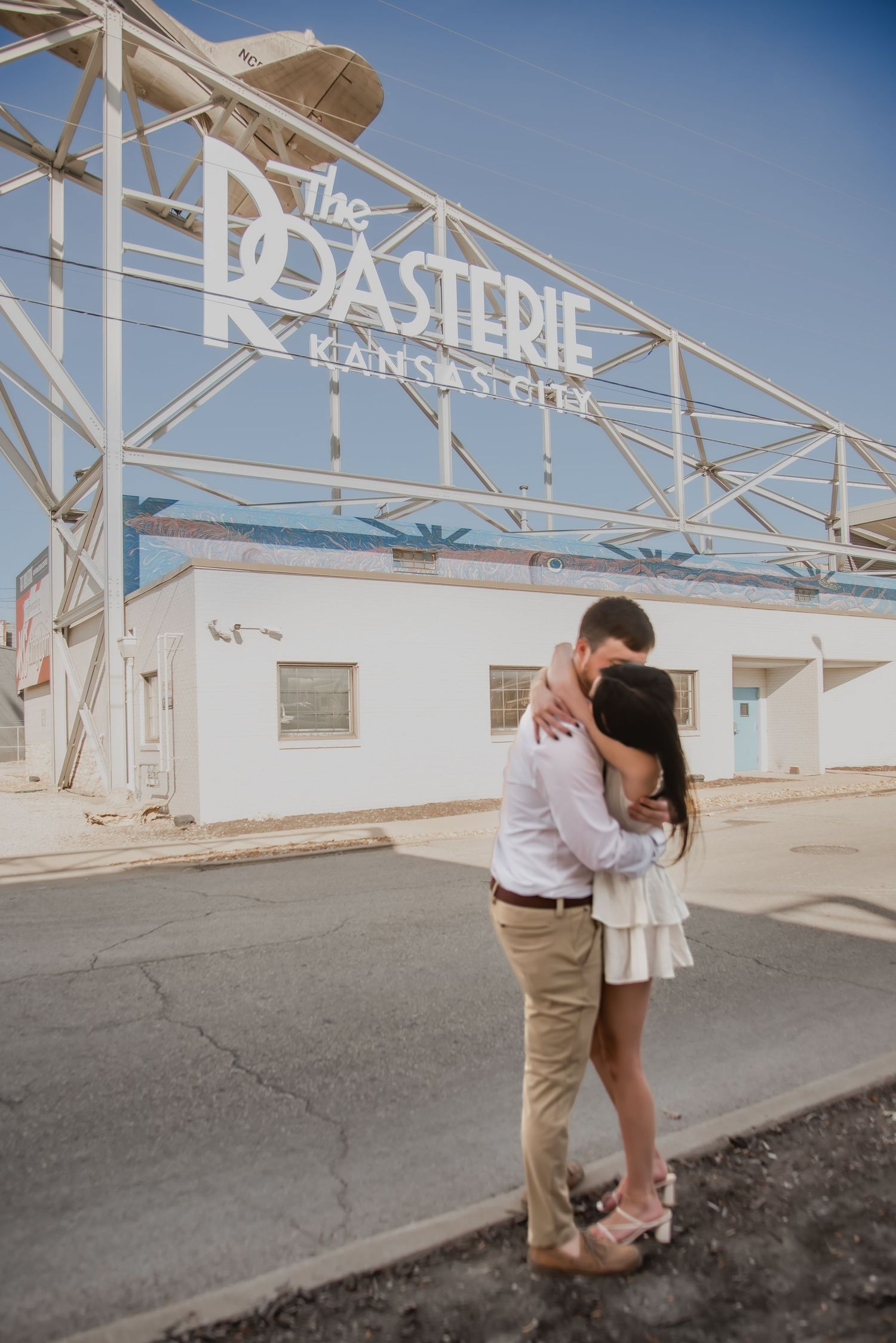 A couple embraces in front of the vintage Roasterie sign in Kansas City on a sunny day.