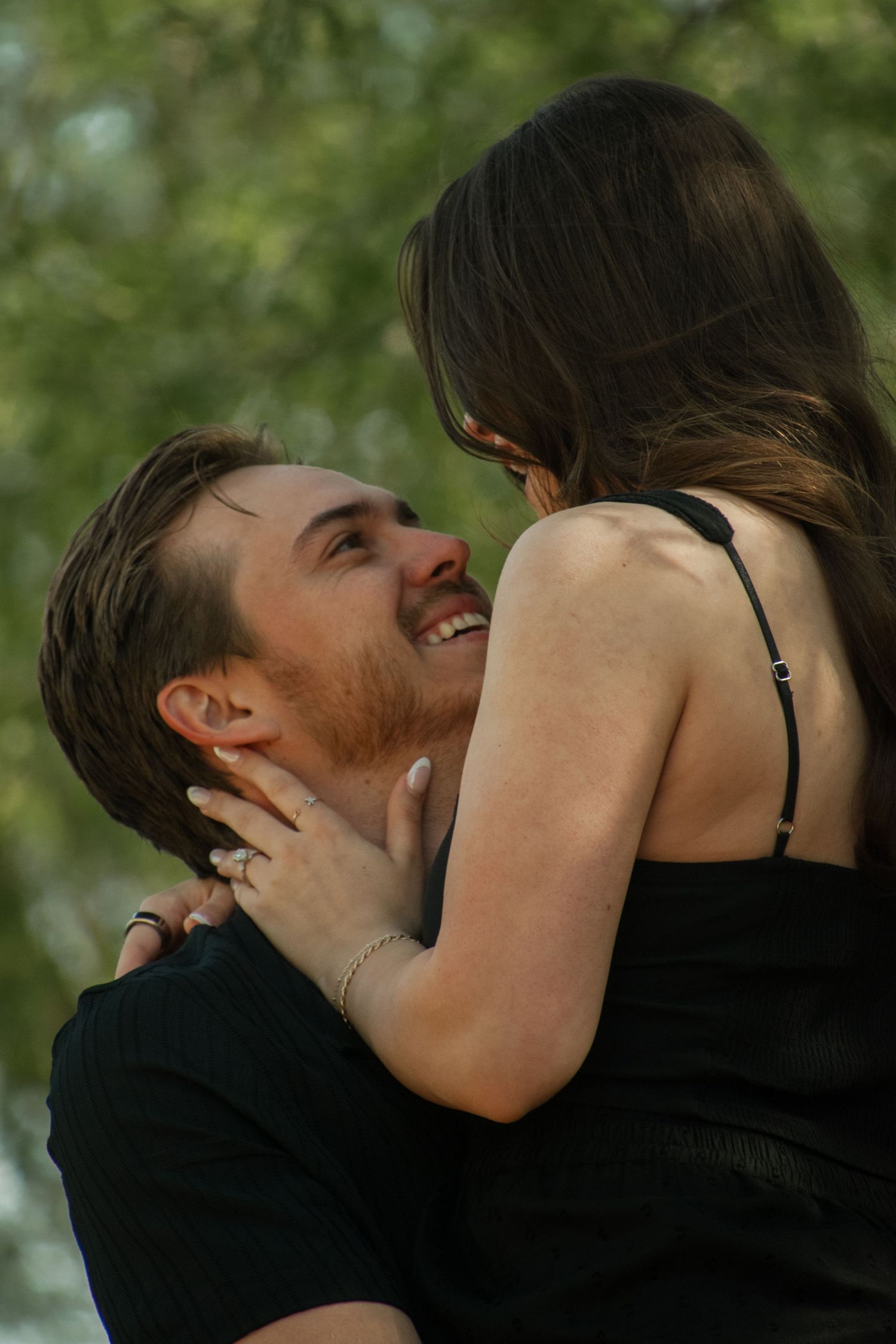 A romantic couple embraces and shares an intimate moment outdoors under green leafy trees.
