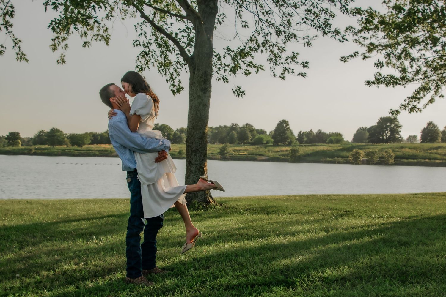 A couple shares a romantic moment on a grassy hillside beneath a towering tree during golden hour.