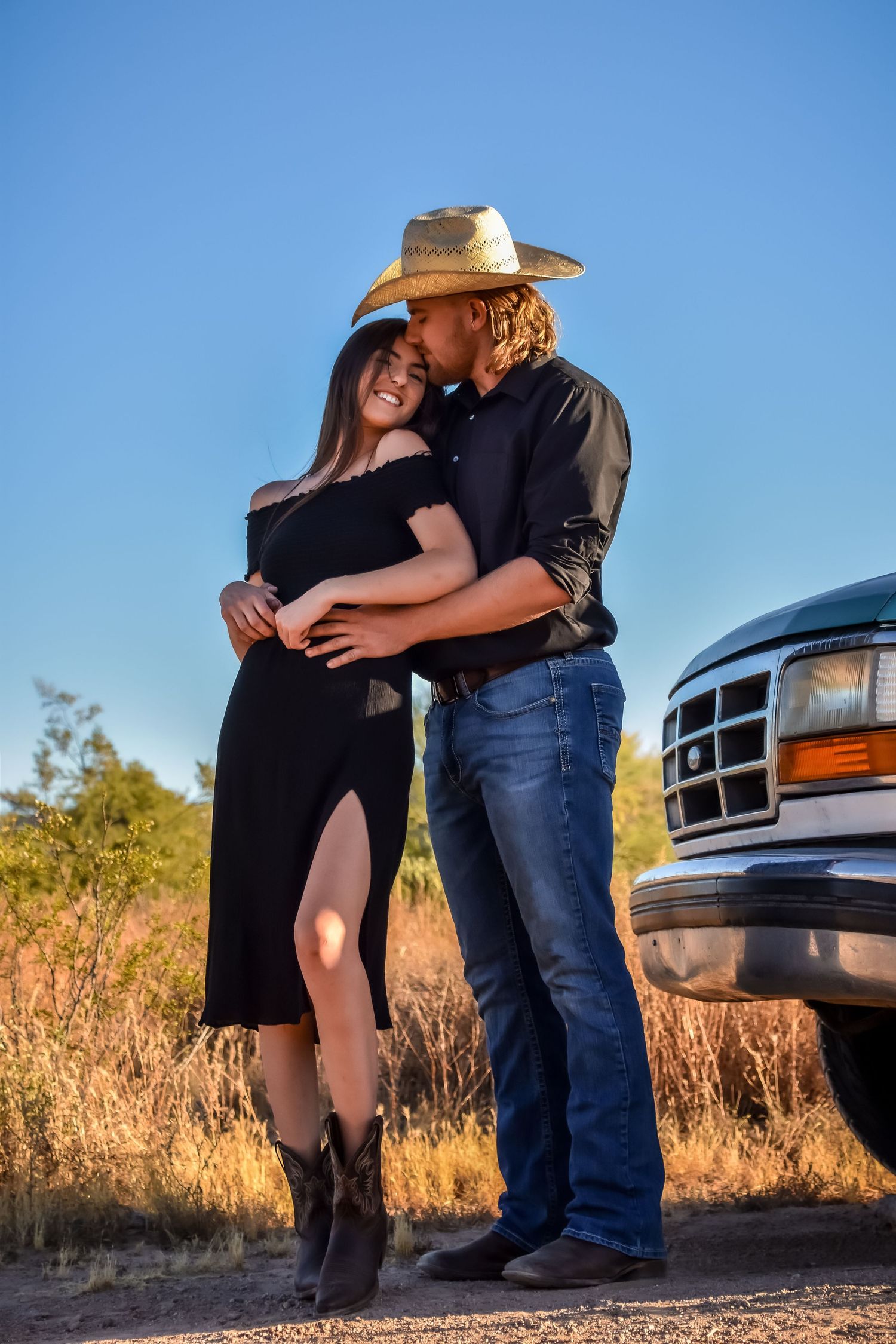 A romantic couple embraces against a backdrop of trucks and dry grass on a sunny day.