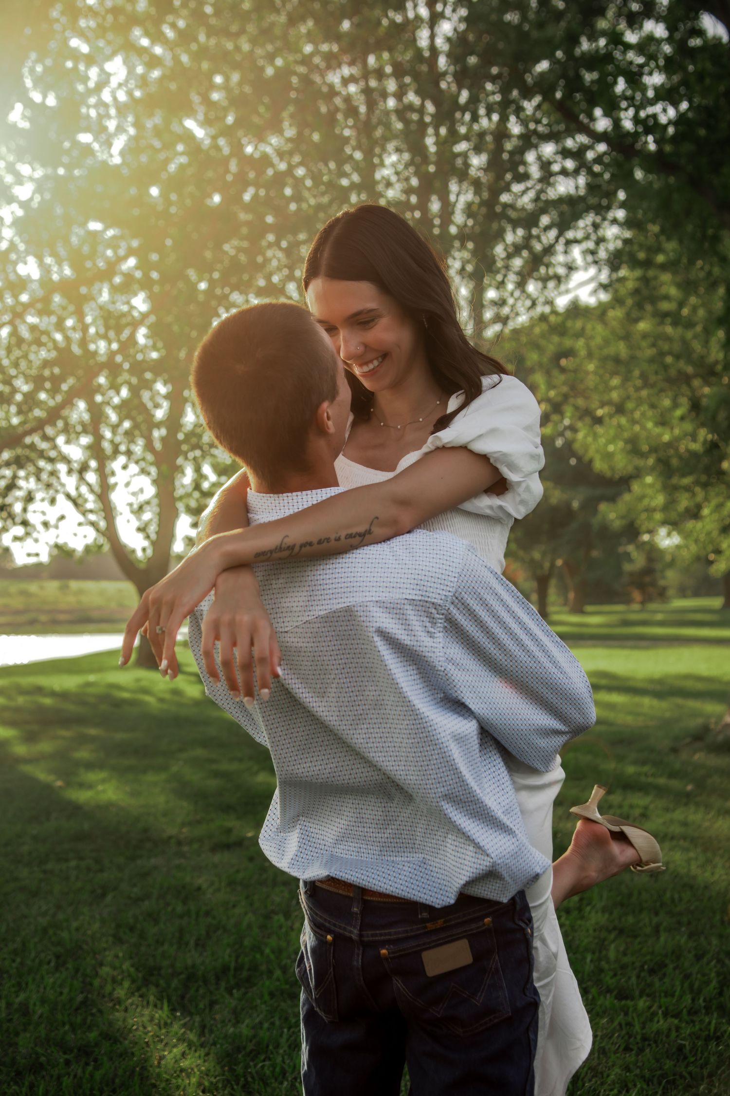 A joyful couple shares a playful embrace on a sunlit lawn beside a lake.