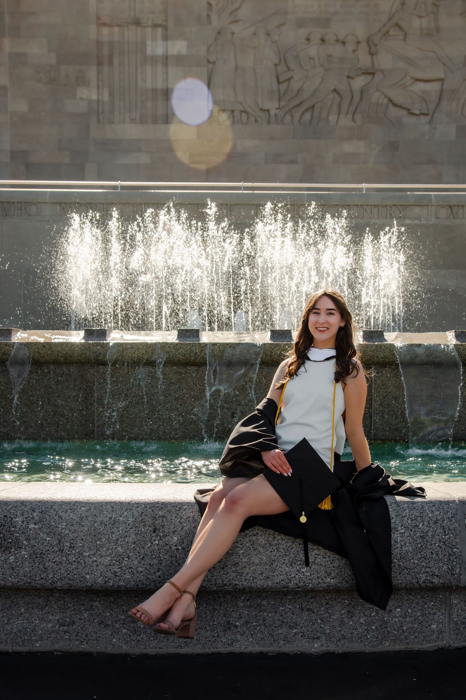 Graduate posing by a fountain wearing academic regalia and white dress for graduation photos.