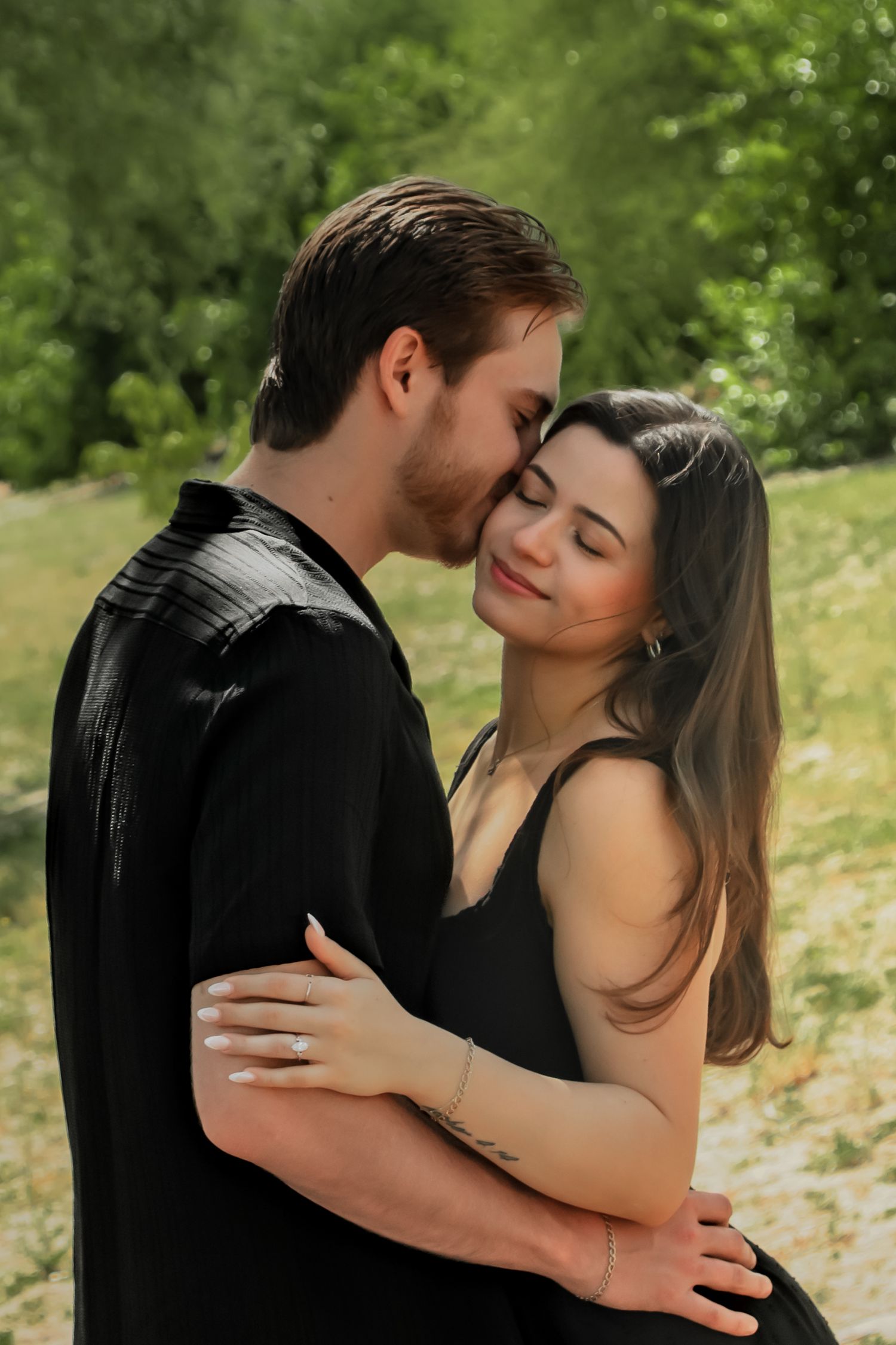Romantic couple in black outfits share an intimate moment outdoors with lush greenery in the background.
