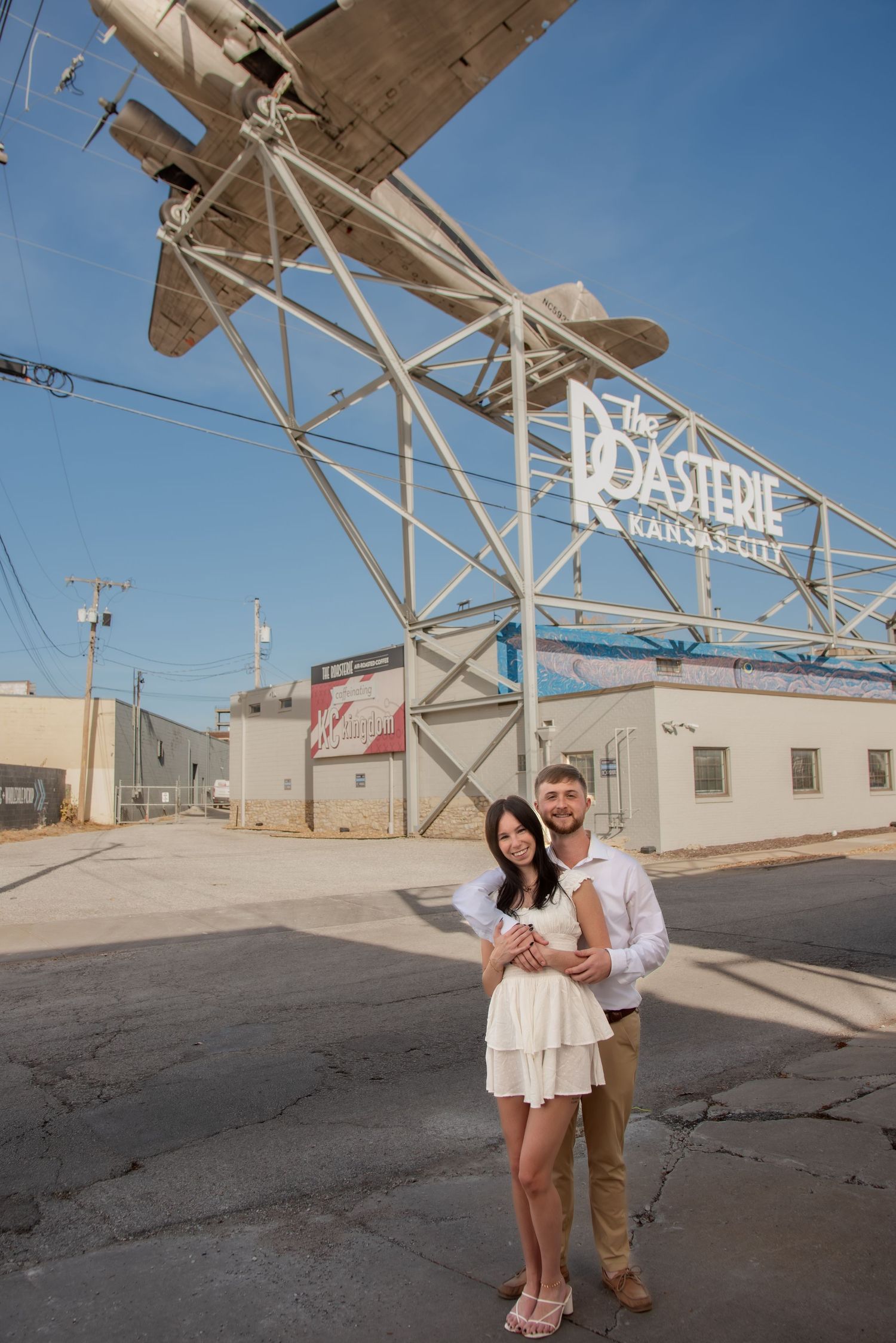 A romantic couple poses in front of an industrial building with white walls and metal framework on a sunny day.