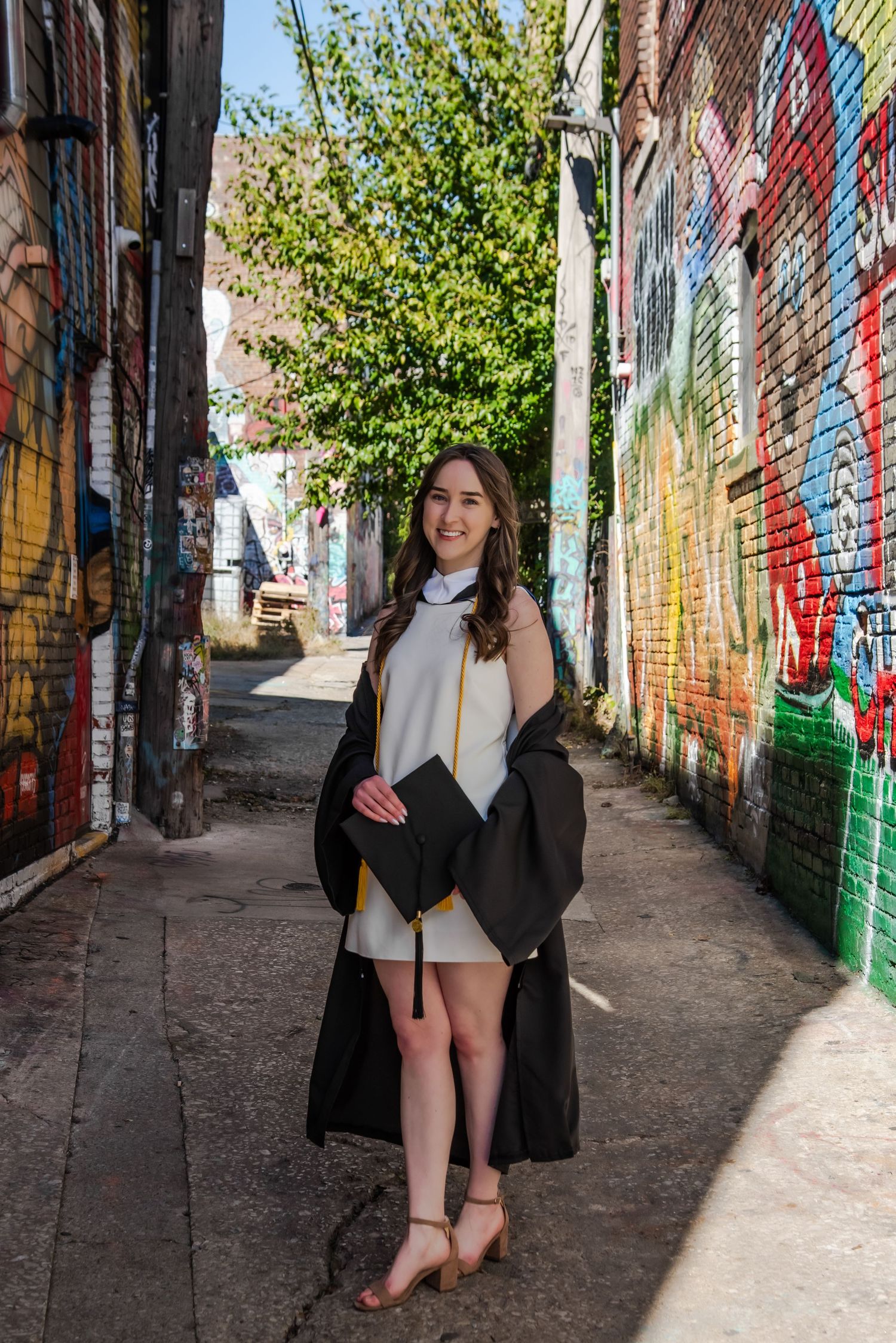 Graduate poses in alley with vibrant graffiti-covered walls and greenery.