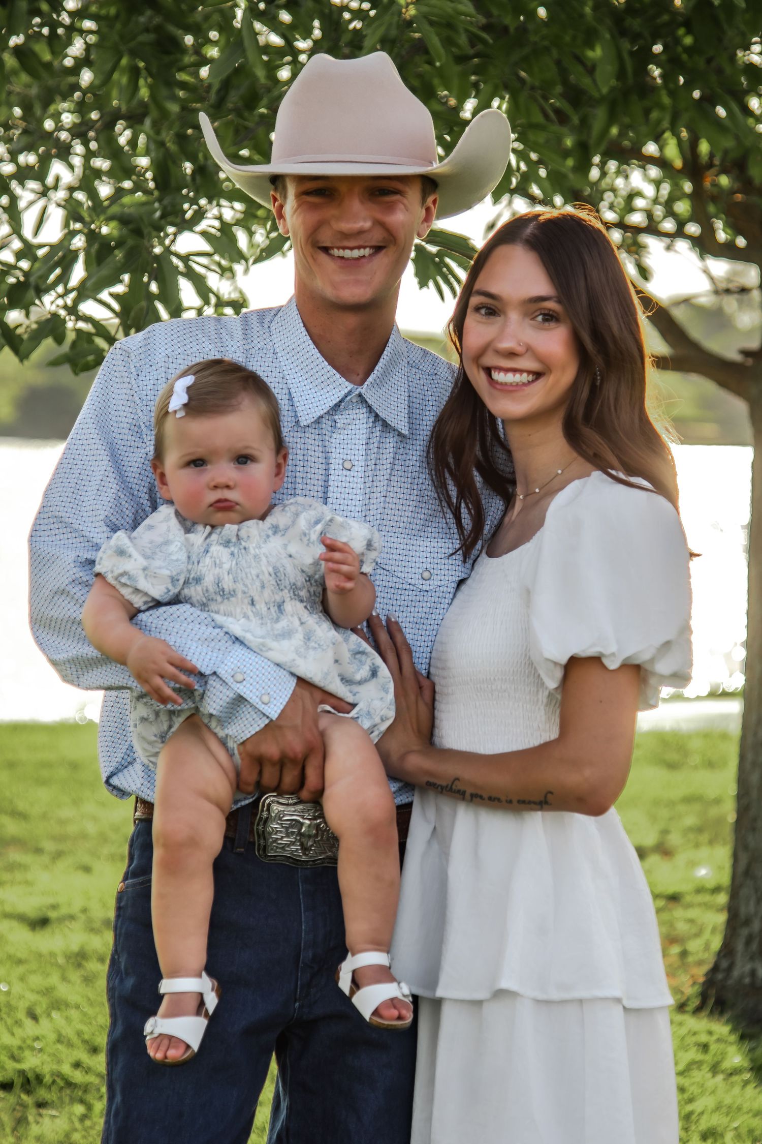 A joyful family poses outdoors together wearing western attire with a baby in blue dress.