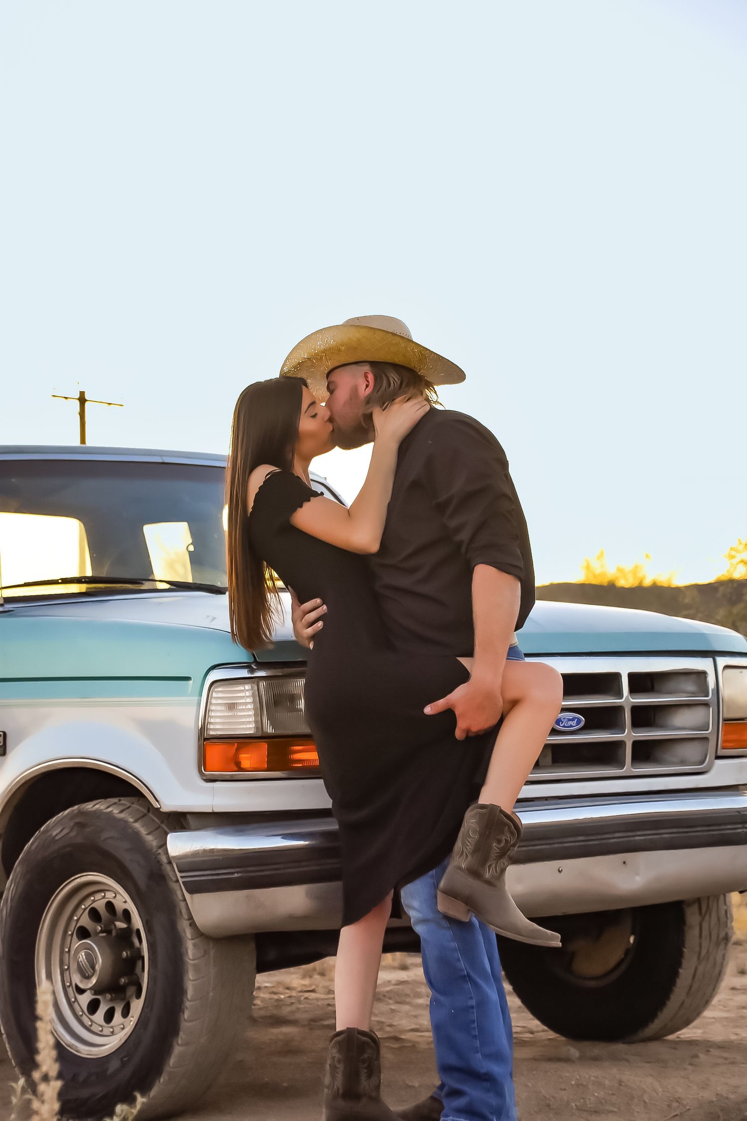 A romantic couple embraces against a pickup truck at sunset in a rustic country setting.