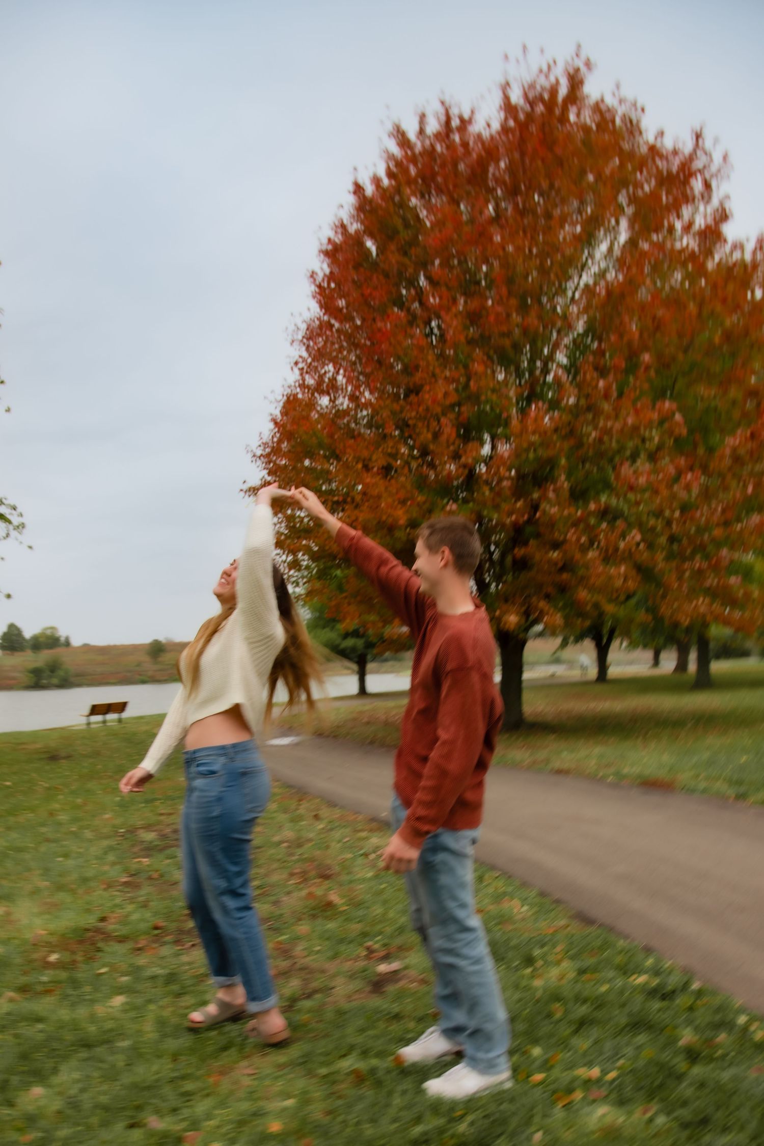 Two people playfully dance together on a path surrounded by autumn trees with vibrant orange foliage.