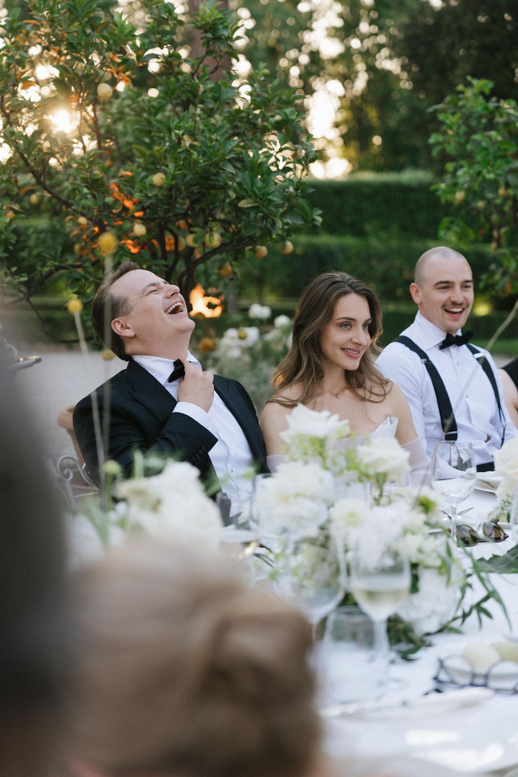 Bride and groom laughing at outdoor wedding reception table with white floral arrangements and lush green garden backdrop.