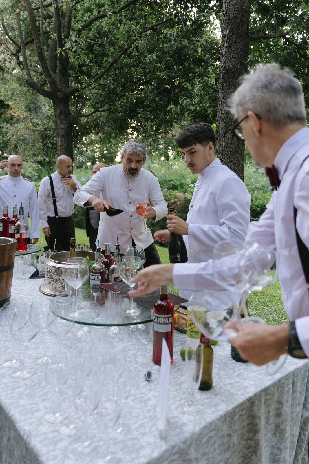 Waiters in white uniforms pour drinks at an outdoor bar table with Campari bottles and wine glasses.