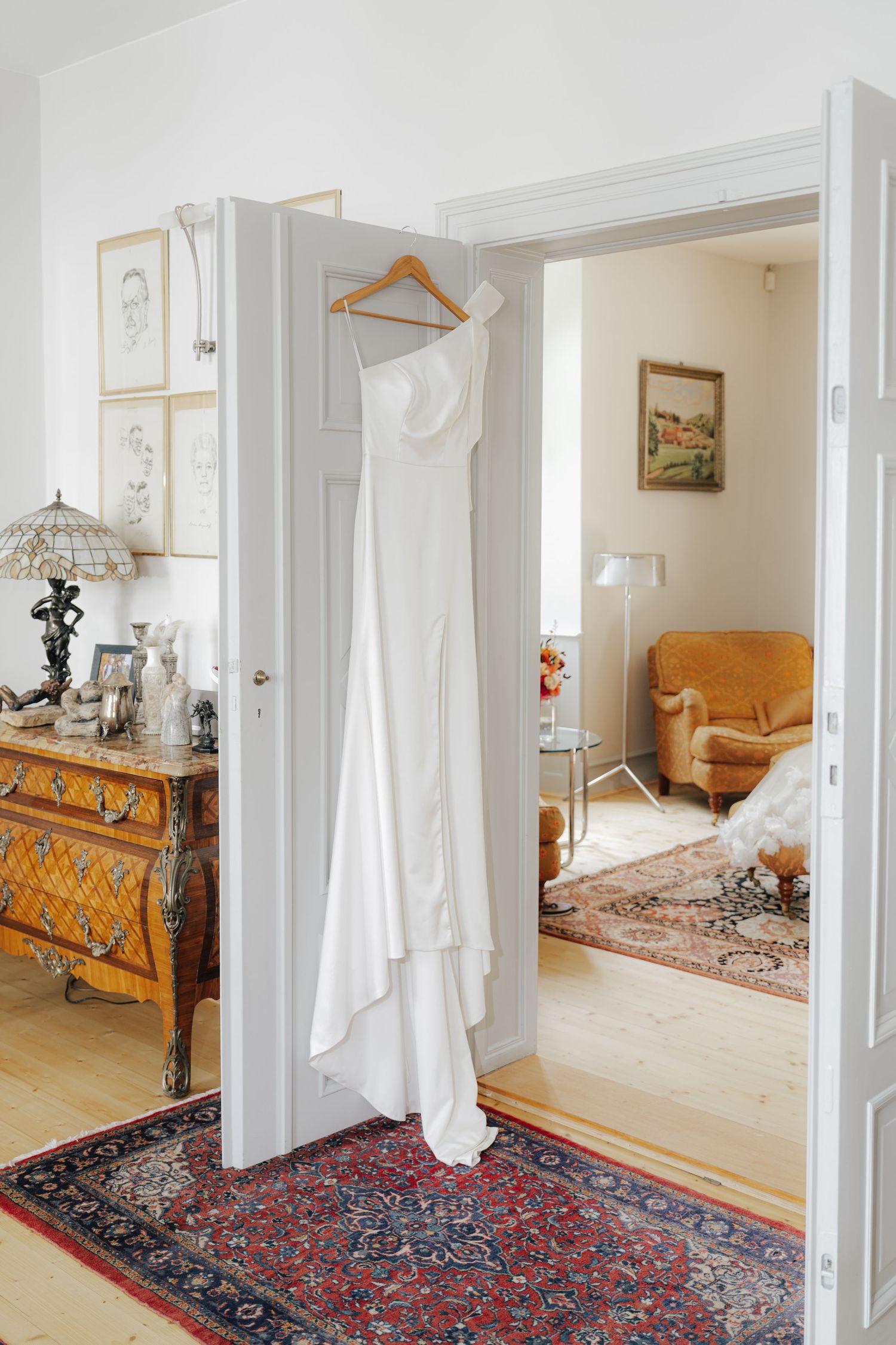 White wedding gown hanging on doorframe in elegant room with antique furniture and oriental rugs.