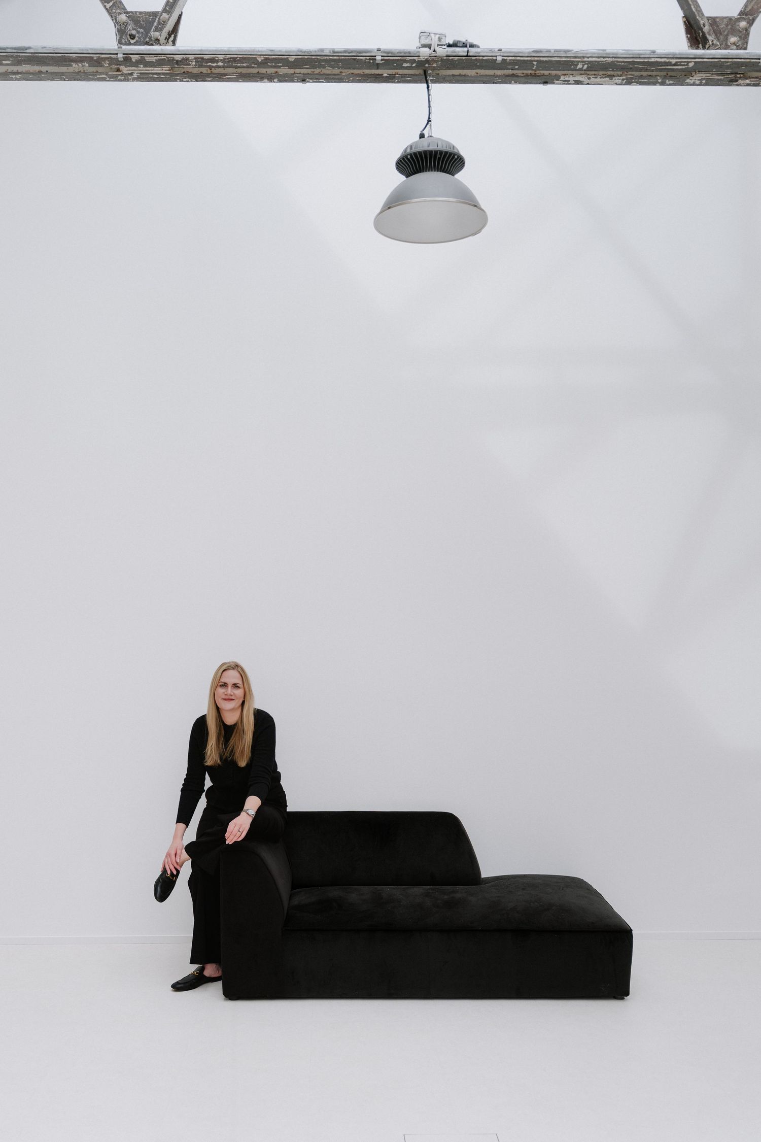 Woman in black outfit sitting on a dark velvet chaise lounge in a minimalist white studio with industrial pendant light.
