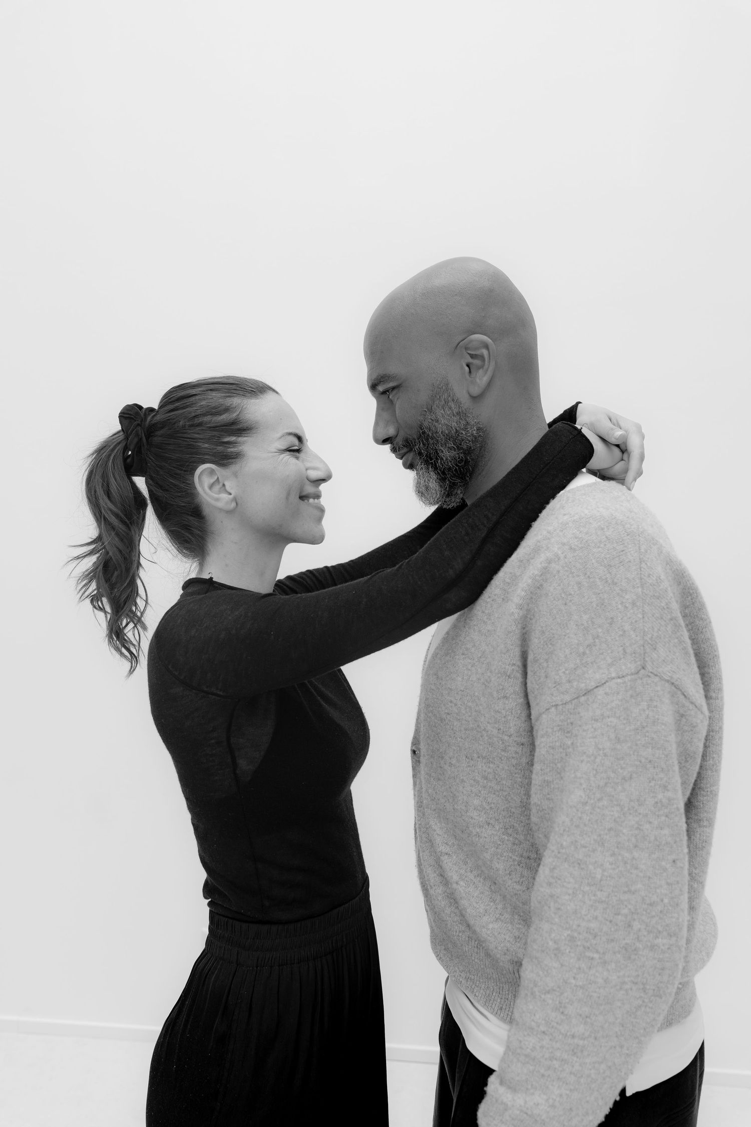 Black and white photo of a smiling couple facing each other, woman in black dress with hand on mans shoulder.