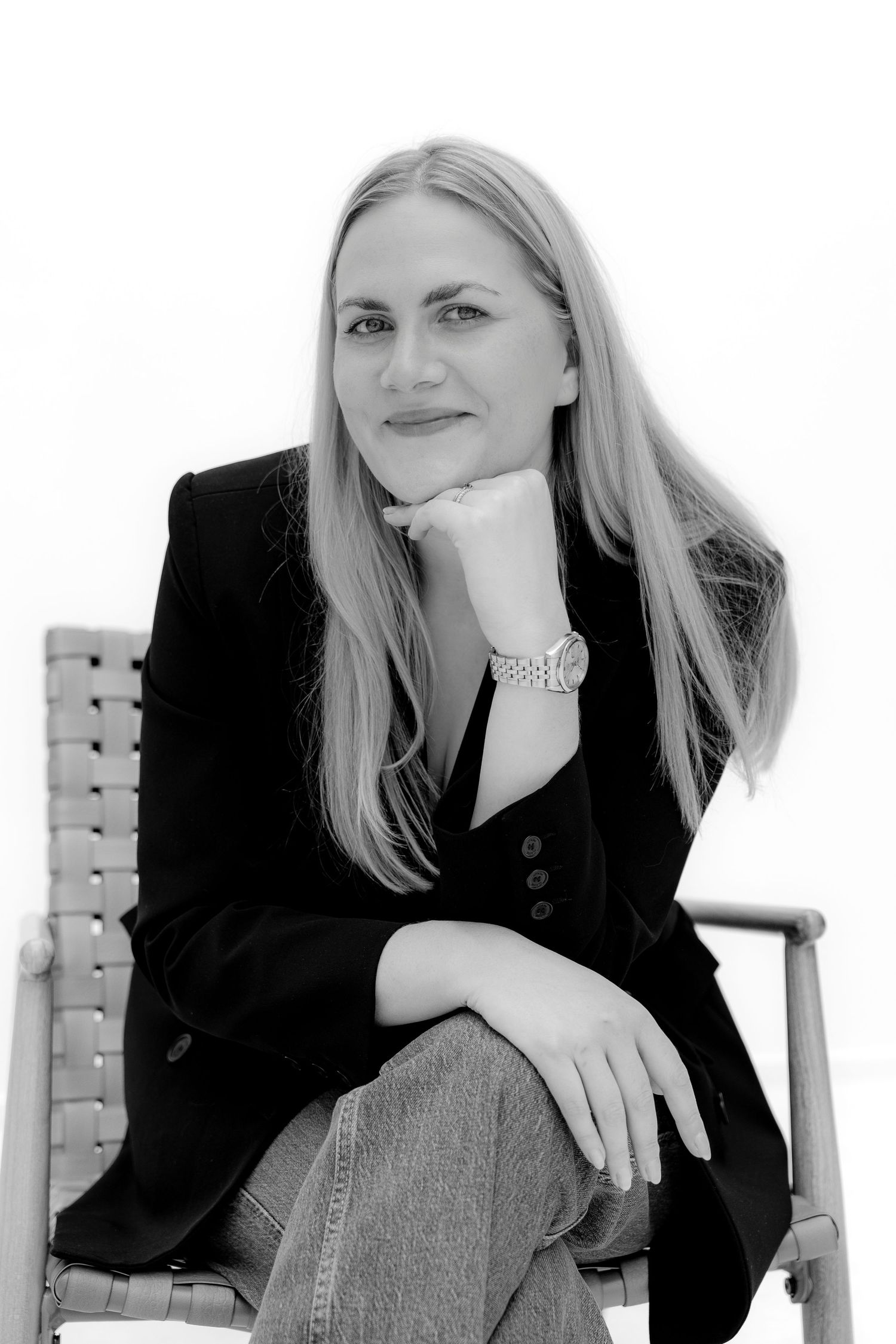 Black and white portrait of a blonde woman in a blazer and jeans, seated in a chair, resting chin on hand.