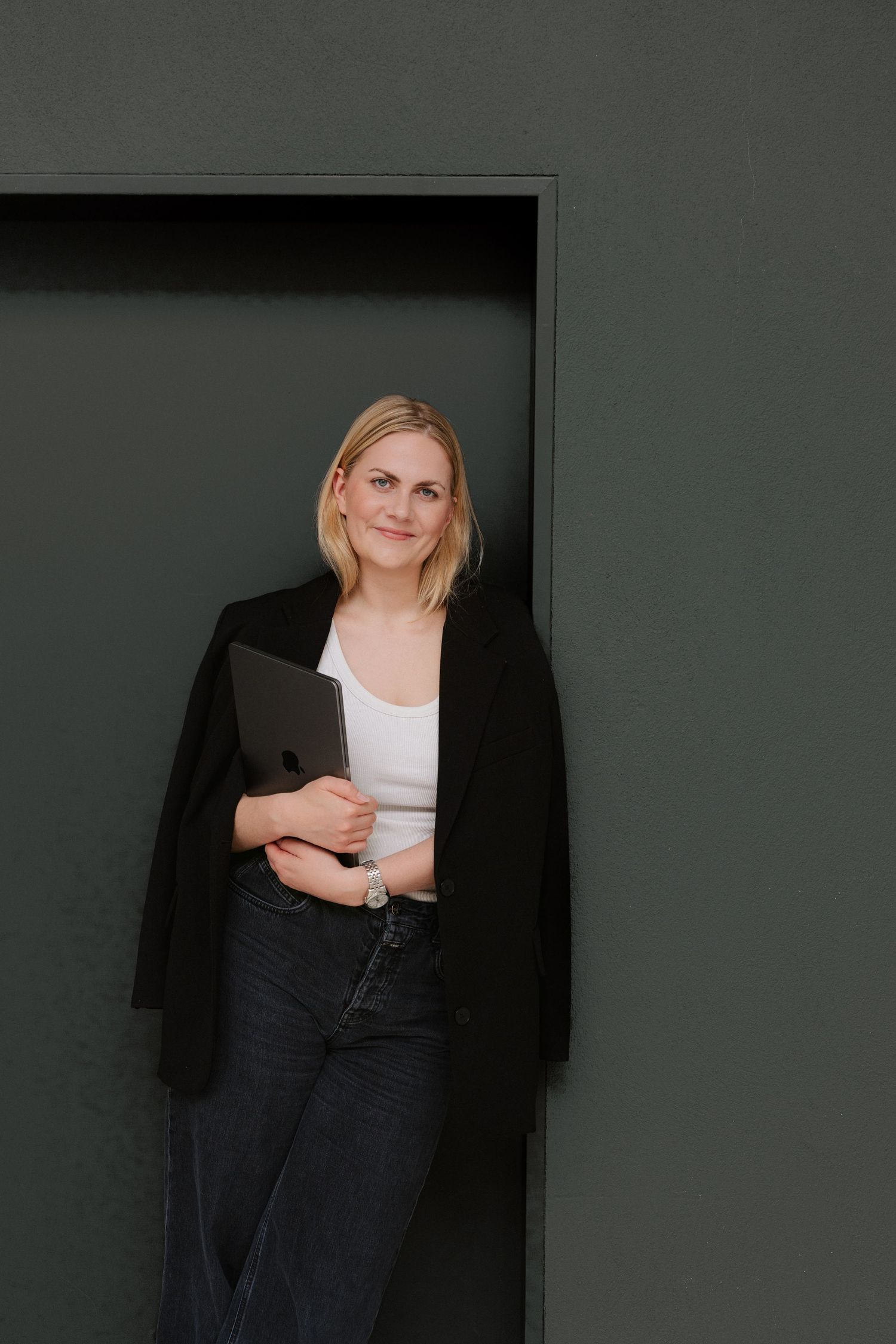 Blonde woman in black blazer and jeans holding a tablet, leaning against a dark green wall.