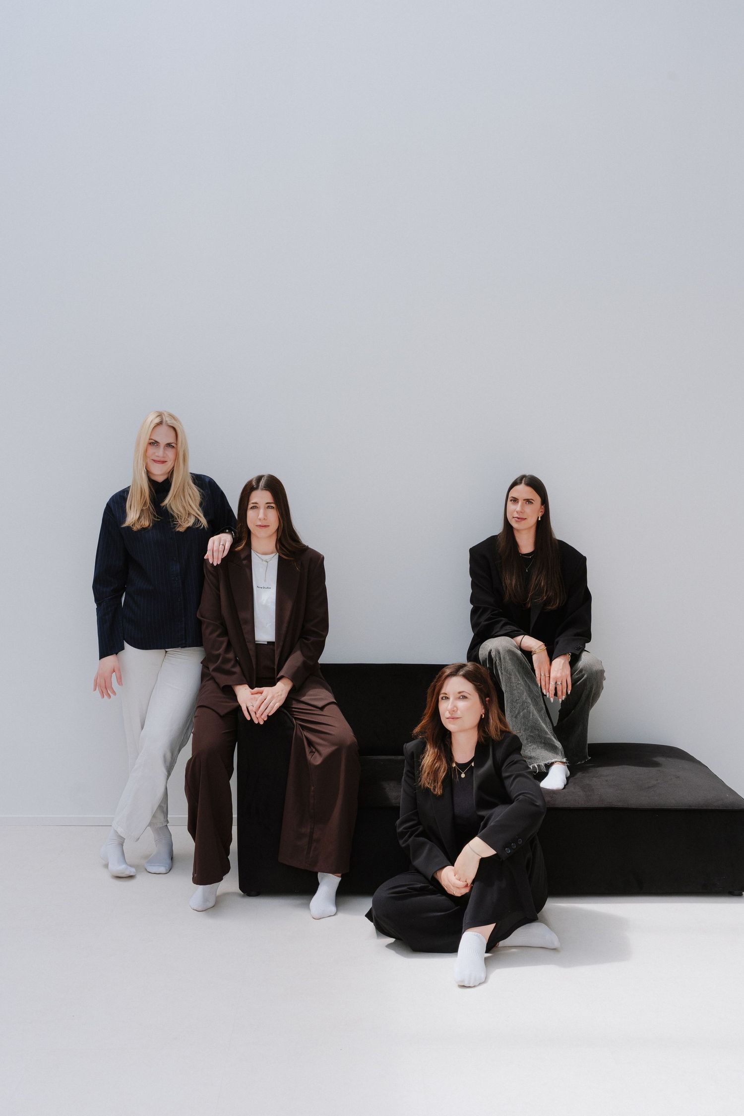 Four women in dark blazers pose against a gray wall, three seated on a bench and one on the floor.