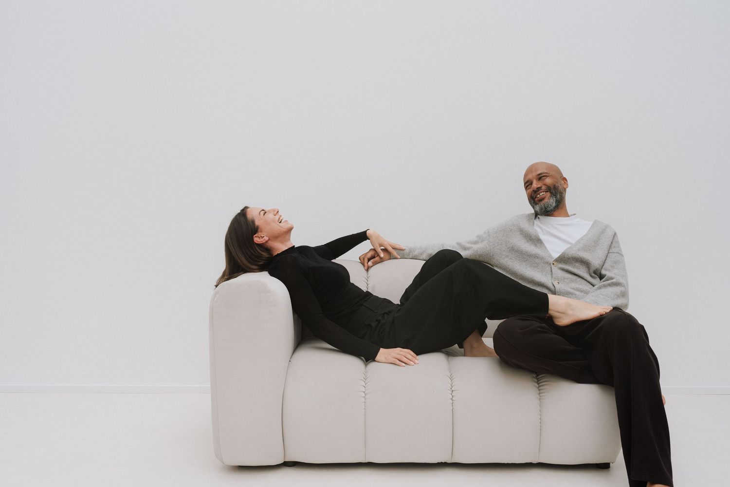 Man and woman relaxing on a modern white sofa against a light gray background in casual clothing.