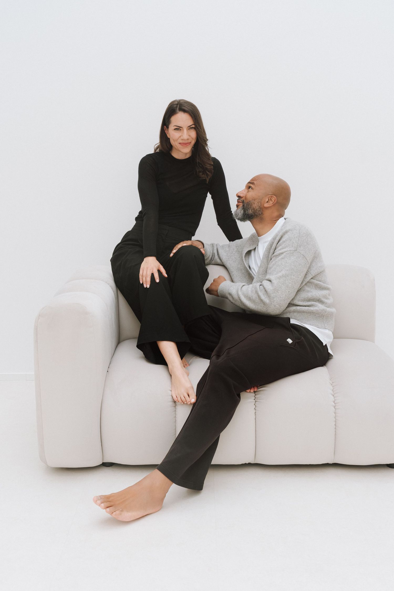 A man and woman sitting together on a white sofa, smiling at each other in a bright, minimalist setting.