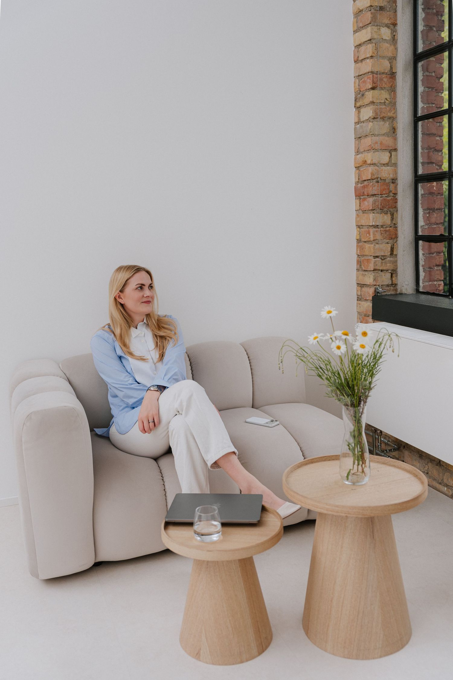 Woman in blue shirt relaxing on beige sofa beside wooden tables with daisies and laptop in modern interior.