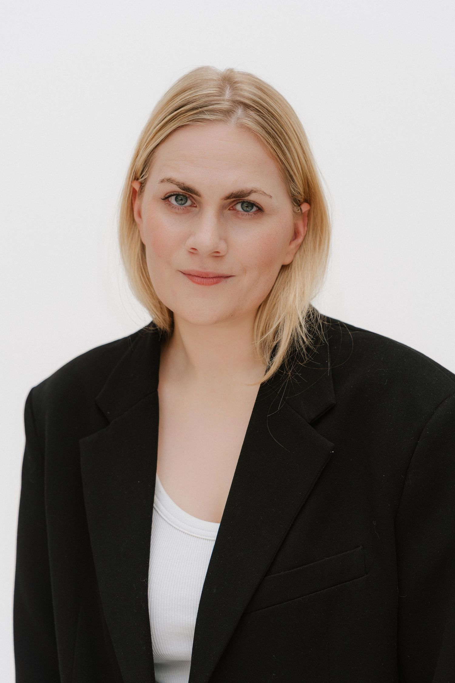 Blonde woman in black blazer and white top poses against white background for professional headshot.