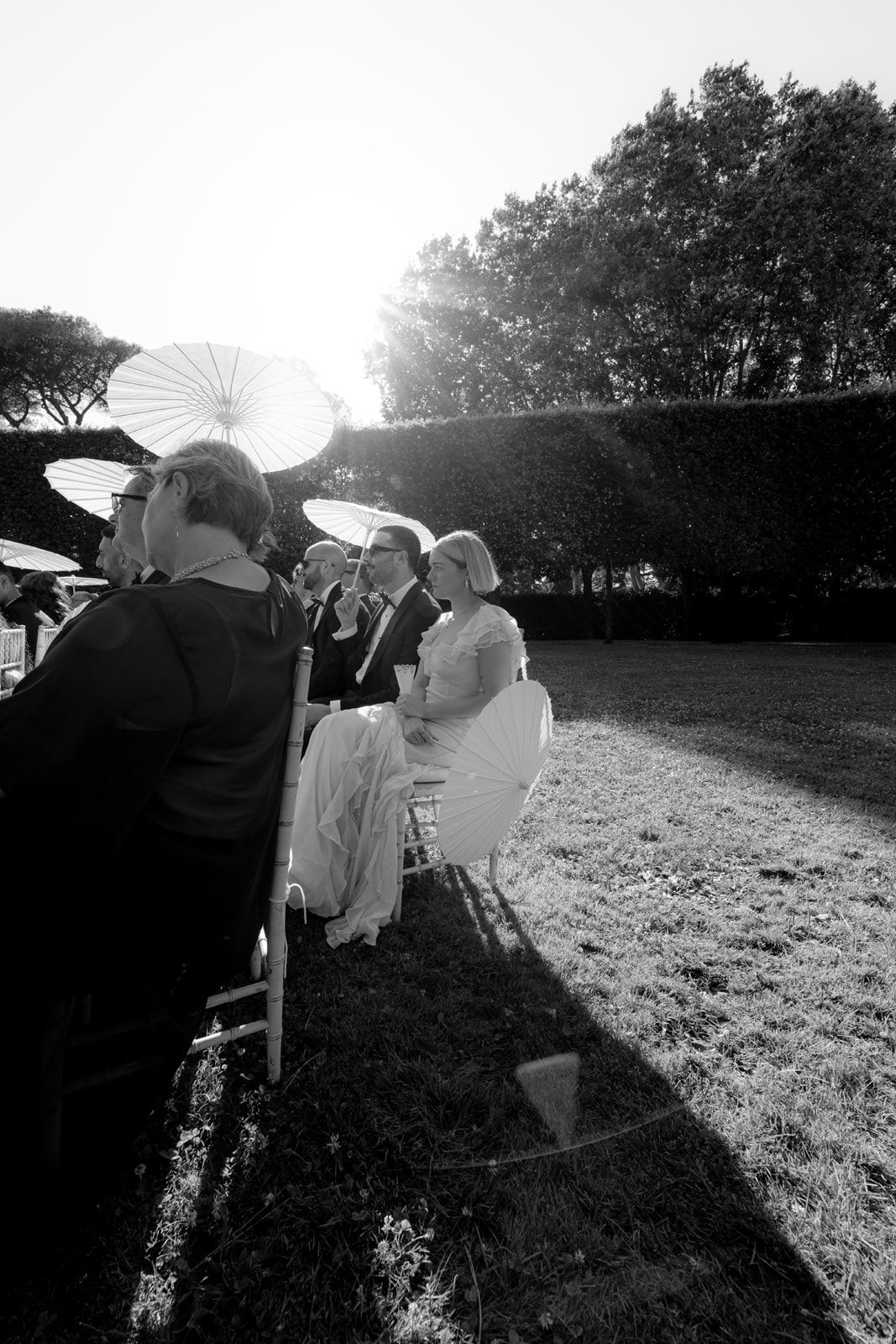 Black and white outdoor wedding ceremony with guests holding parasols seated on chairs in a sunlit garden.