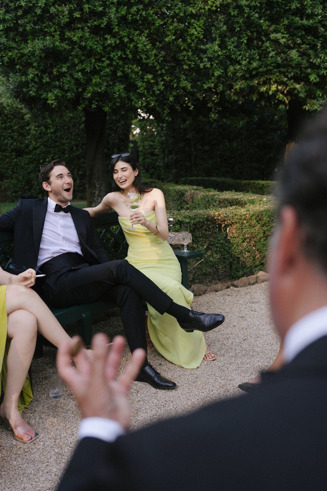 Elegantly dressed couple sitting outdoors at a garden party, woman in yellow gown holding a drink, man in tuxedo.