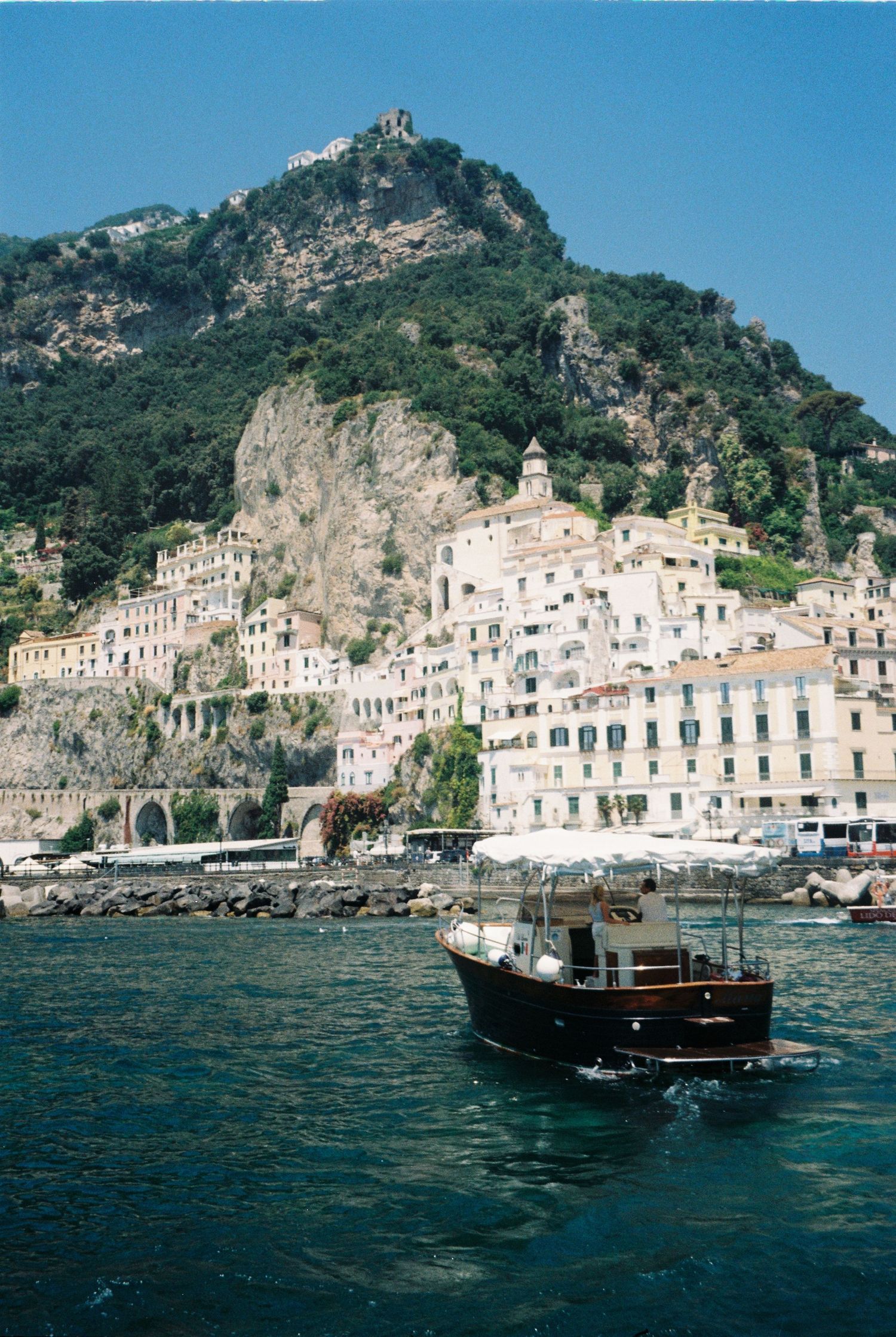 A wooden boat cruises the blue waters of Amalfi Coast, Italy, with colorful cliffside buildings and lush green mountains.