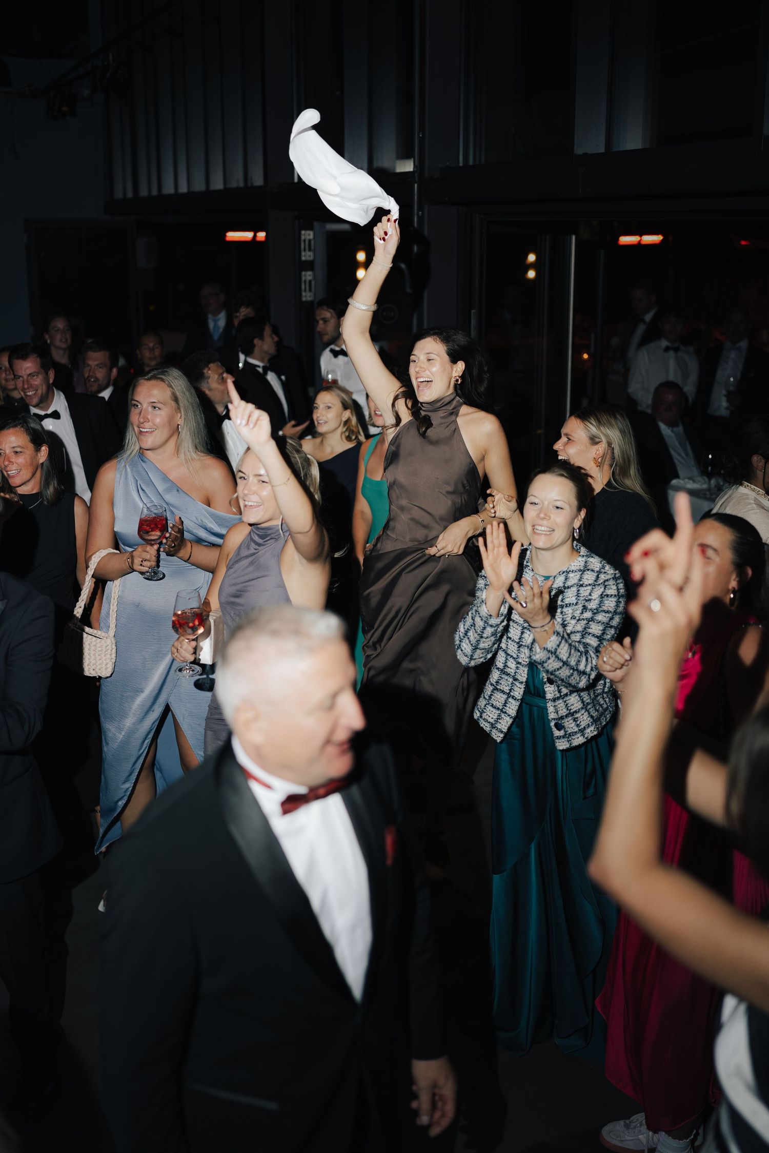 Crowd dancing and waving napkins in the air at a dark reception venue.