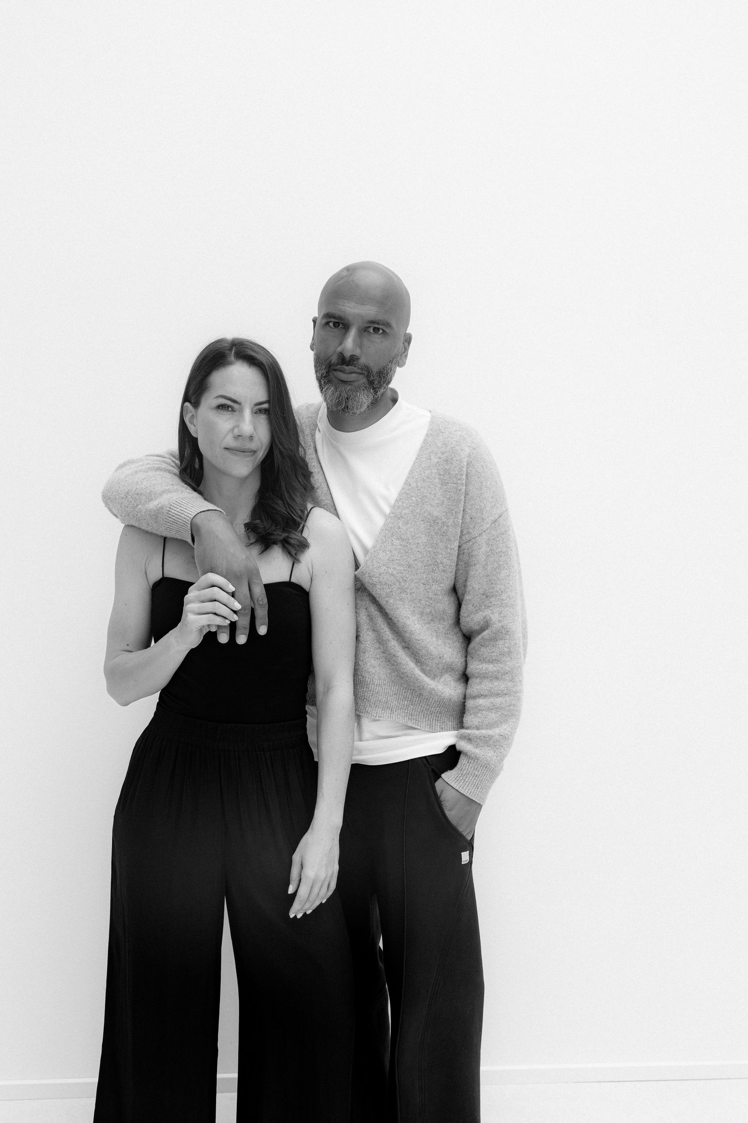 Black and white portrait of a bald man and brunette woman in black outfits posing together against a white wall.