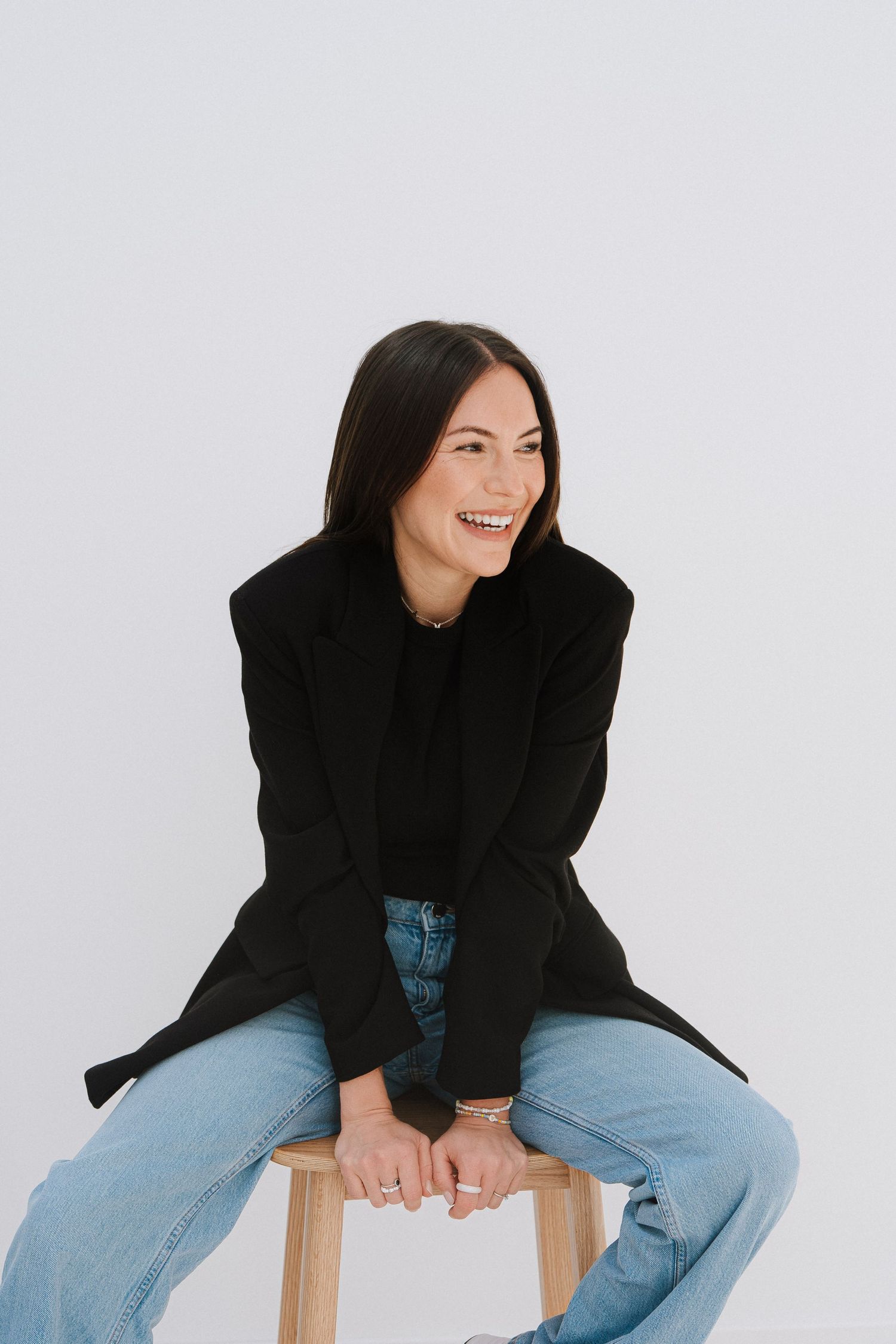 Smiling woman with dark hair in black blazer and jeans sitting on wooden stool against white background.