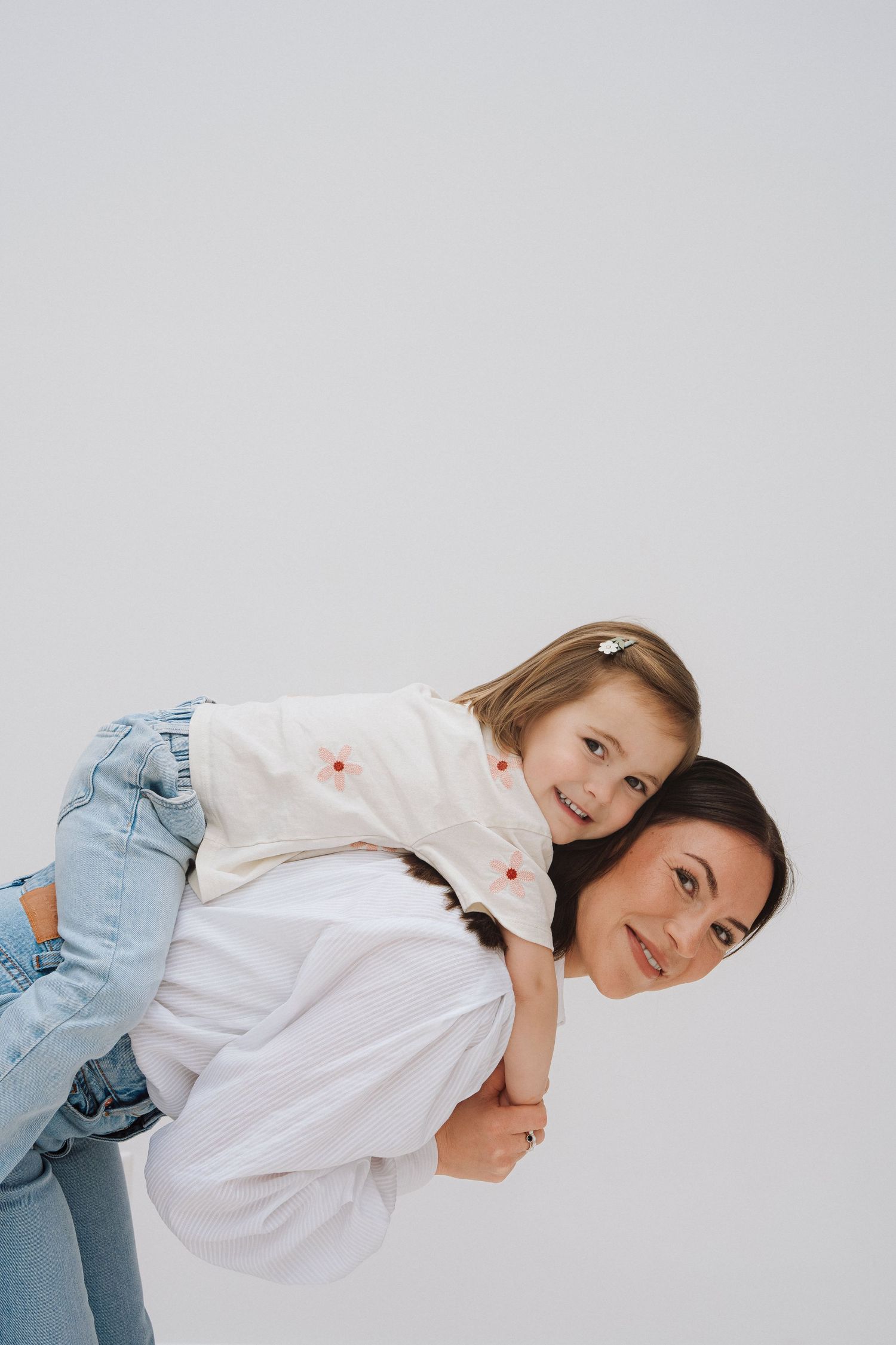 A woman gives a smiling young girl a piggyback ride against a light gray background, both wearing white tops and jeans.