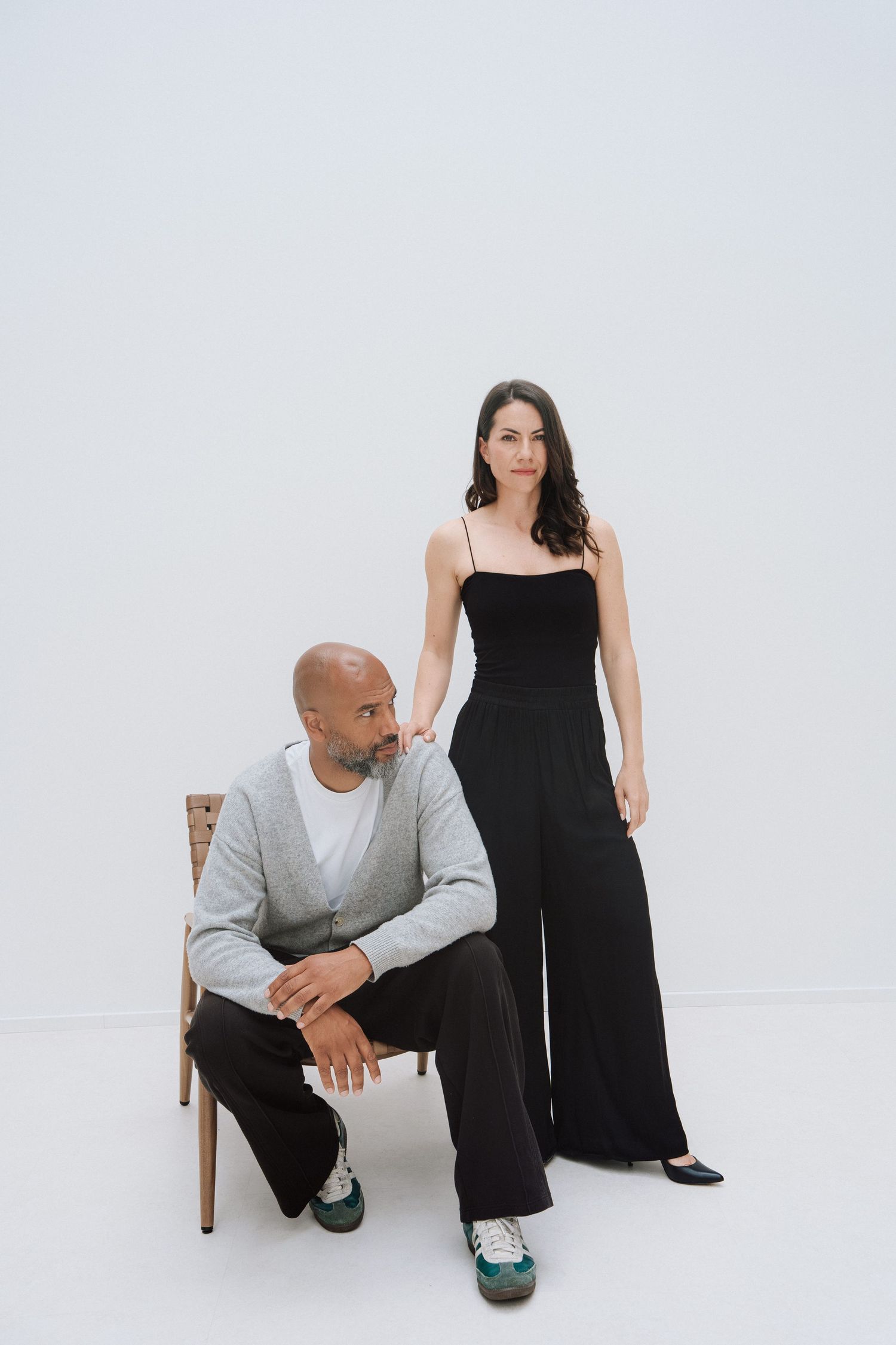 A man sitting on a chair and a woman standing beside him, both wearing black and white outfits against a white background.