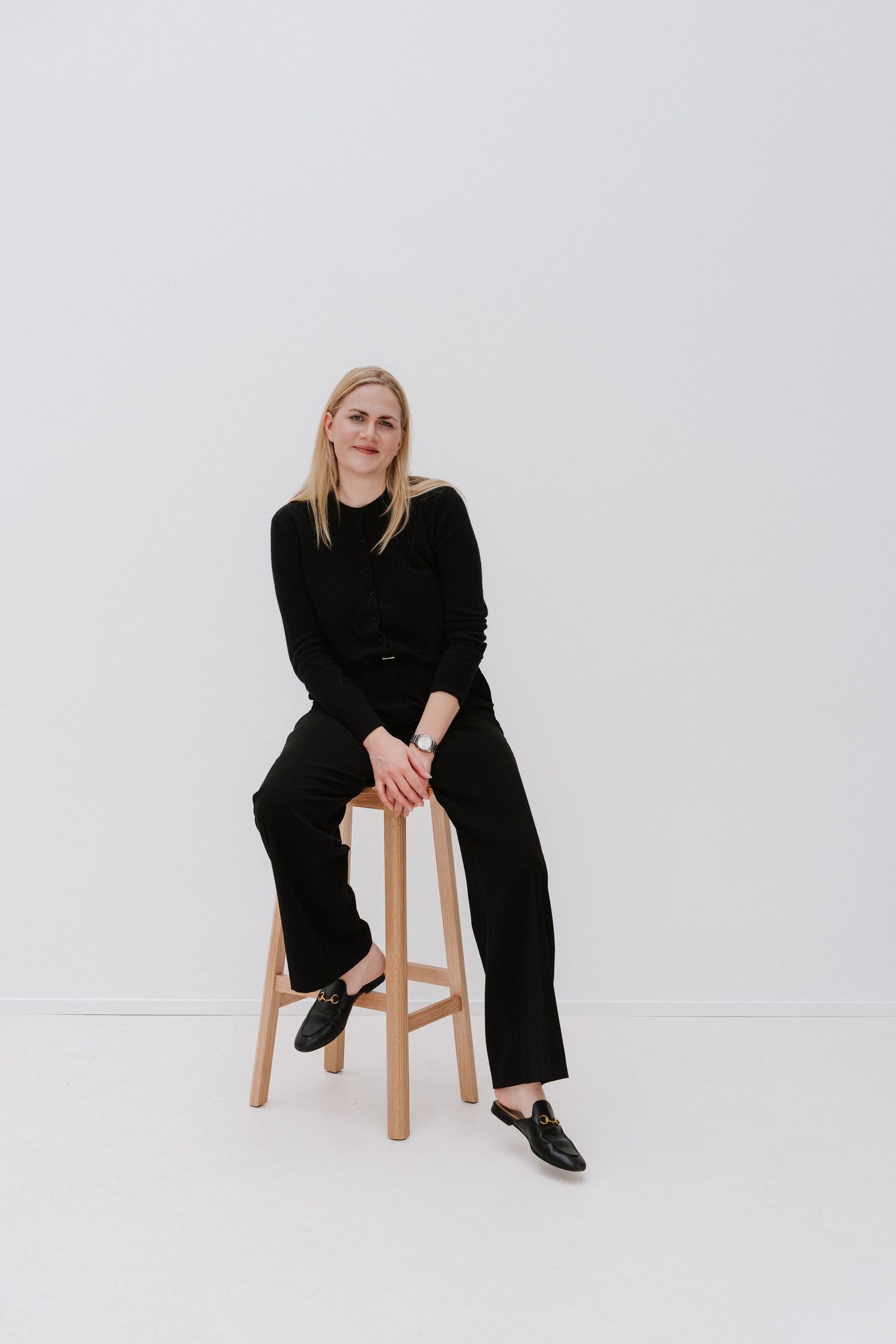 Woman in black outfit sitting on wooden stool against white background, smiling at camera.