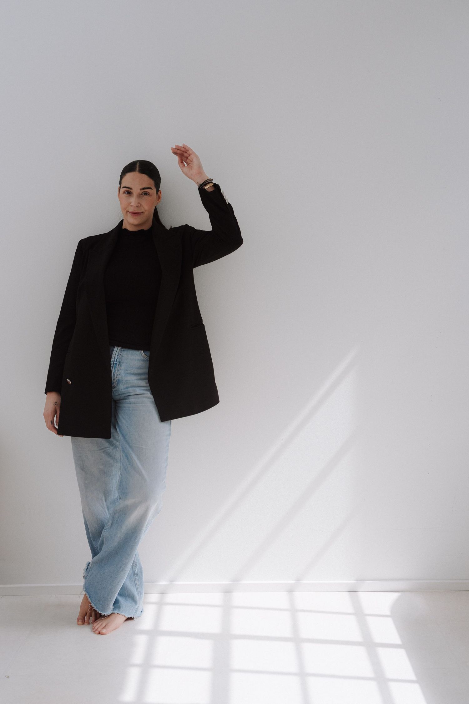Woman in black blazer, turtleneck, and light blue wide-leg jeans posing barefoot against a white wall.