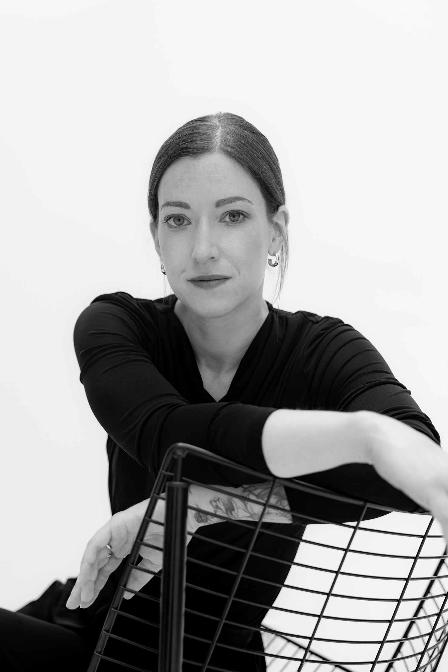 Black and white portrait of a woman in black top leaning on a wire chair, looking confidently at camera.
