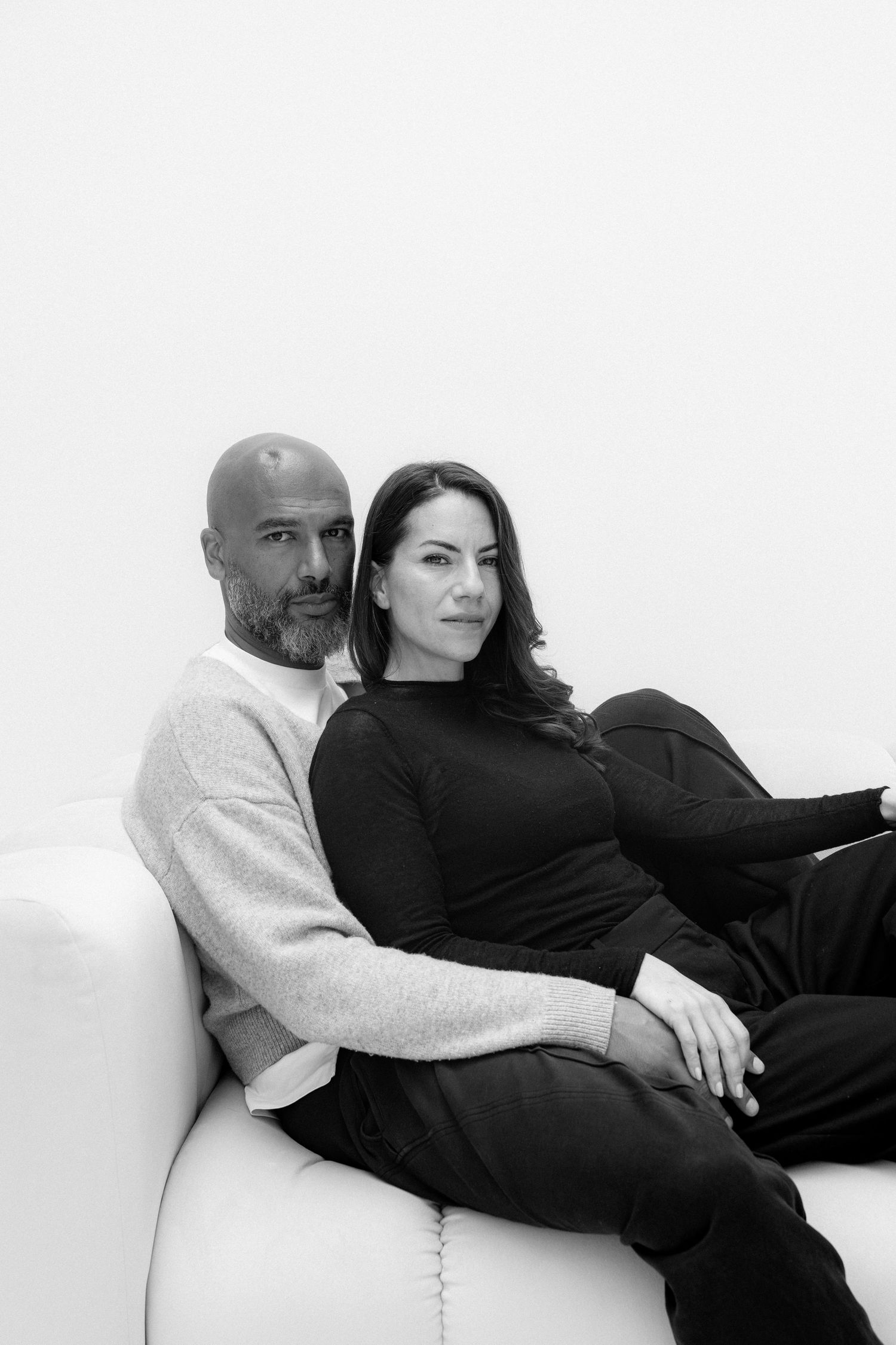 Black and white portrait of a bald man and dark-haired woman sitting together on a white sofa.
