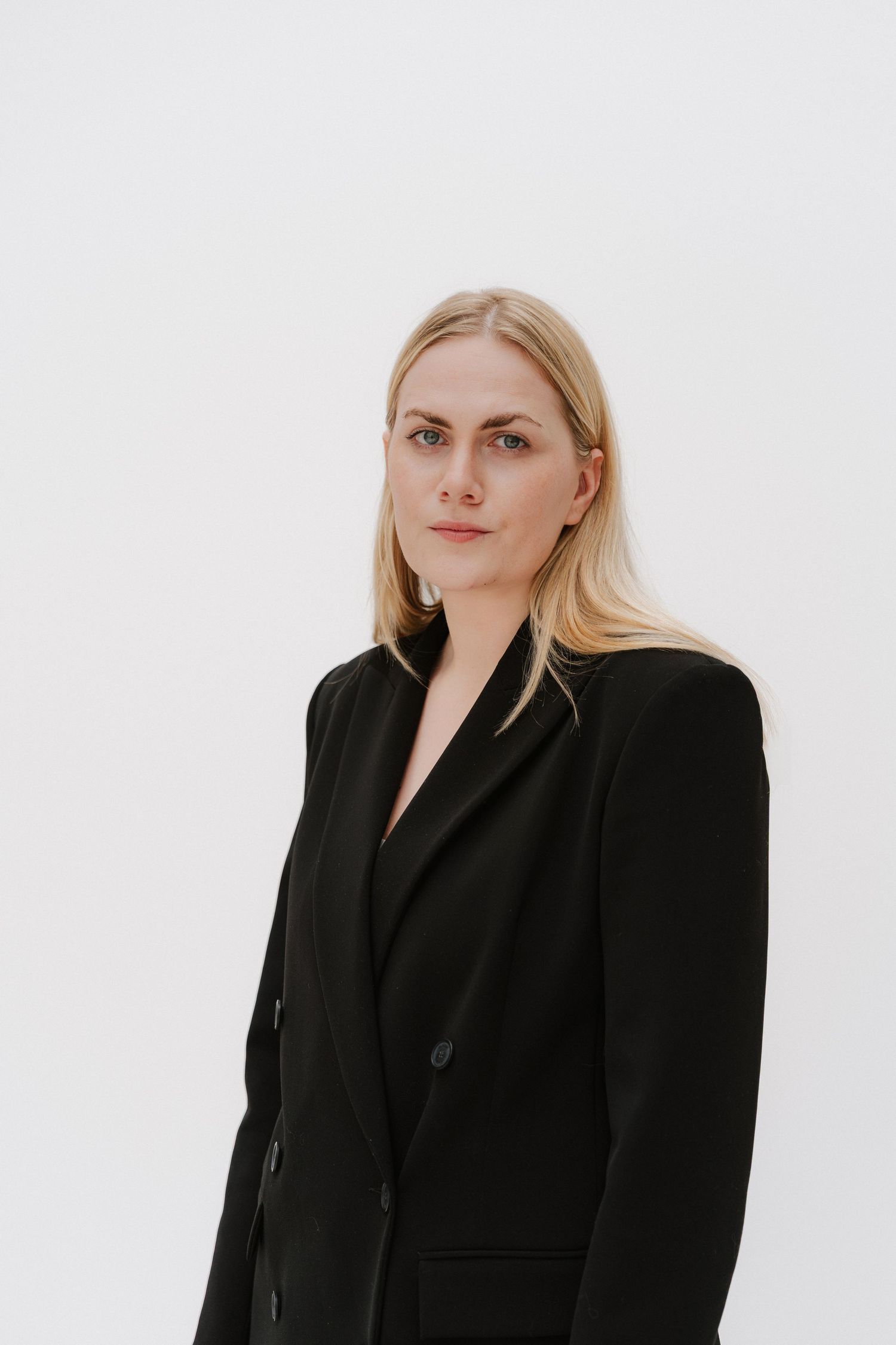 Blonde woman in black blazer poses against white background in professional portrait photo.